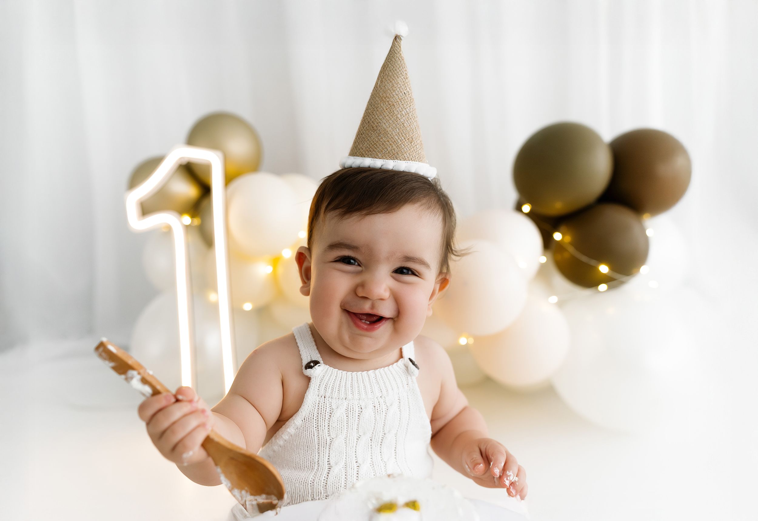 A baby in a white outfit sits on a blanket with gold number one decorations and gold and black balloons.