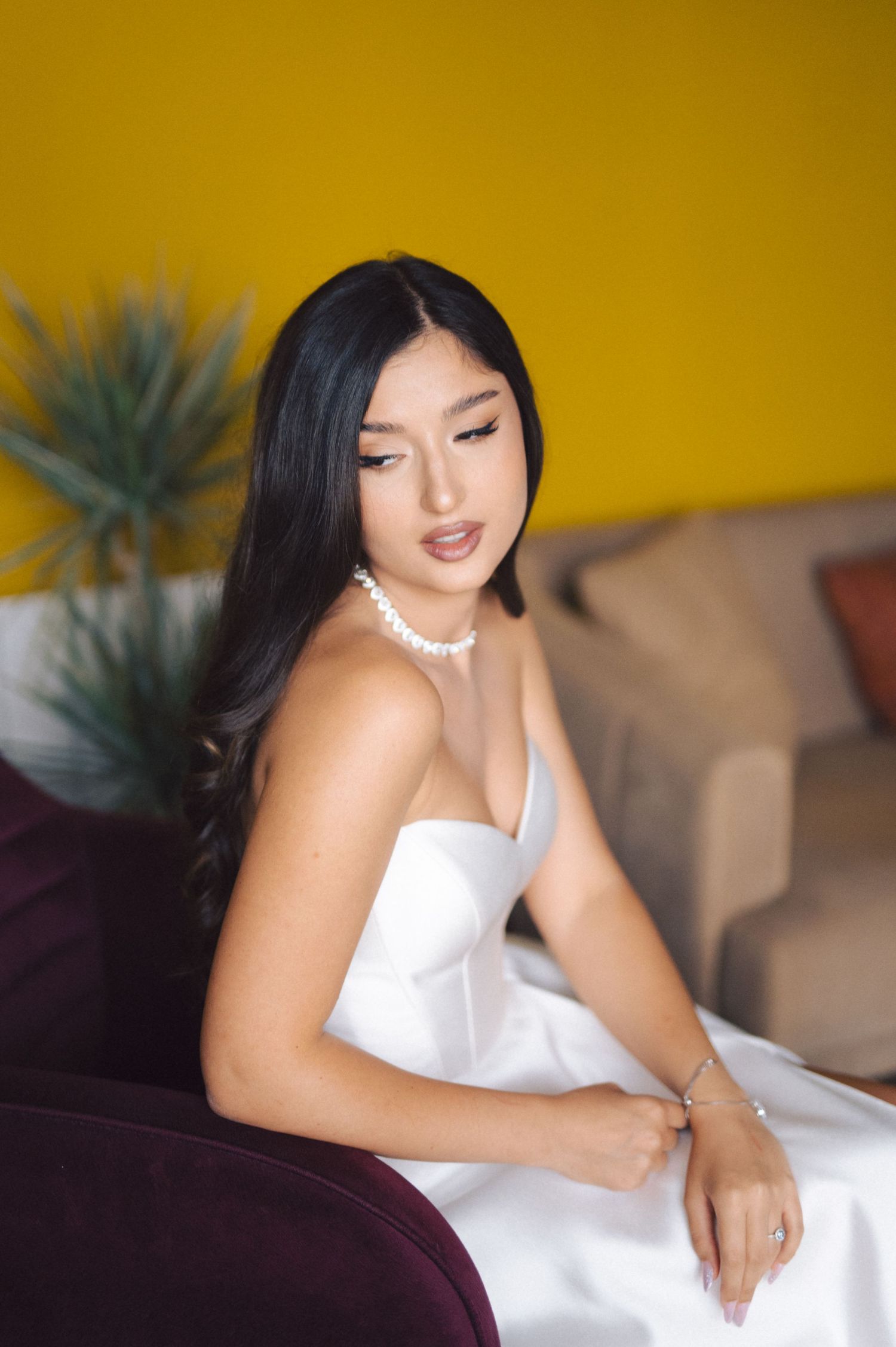 A portrait in a white dress and pearl necklace against a yellow wall with a houseplant in soft focus.