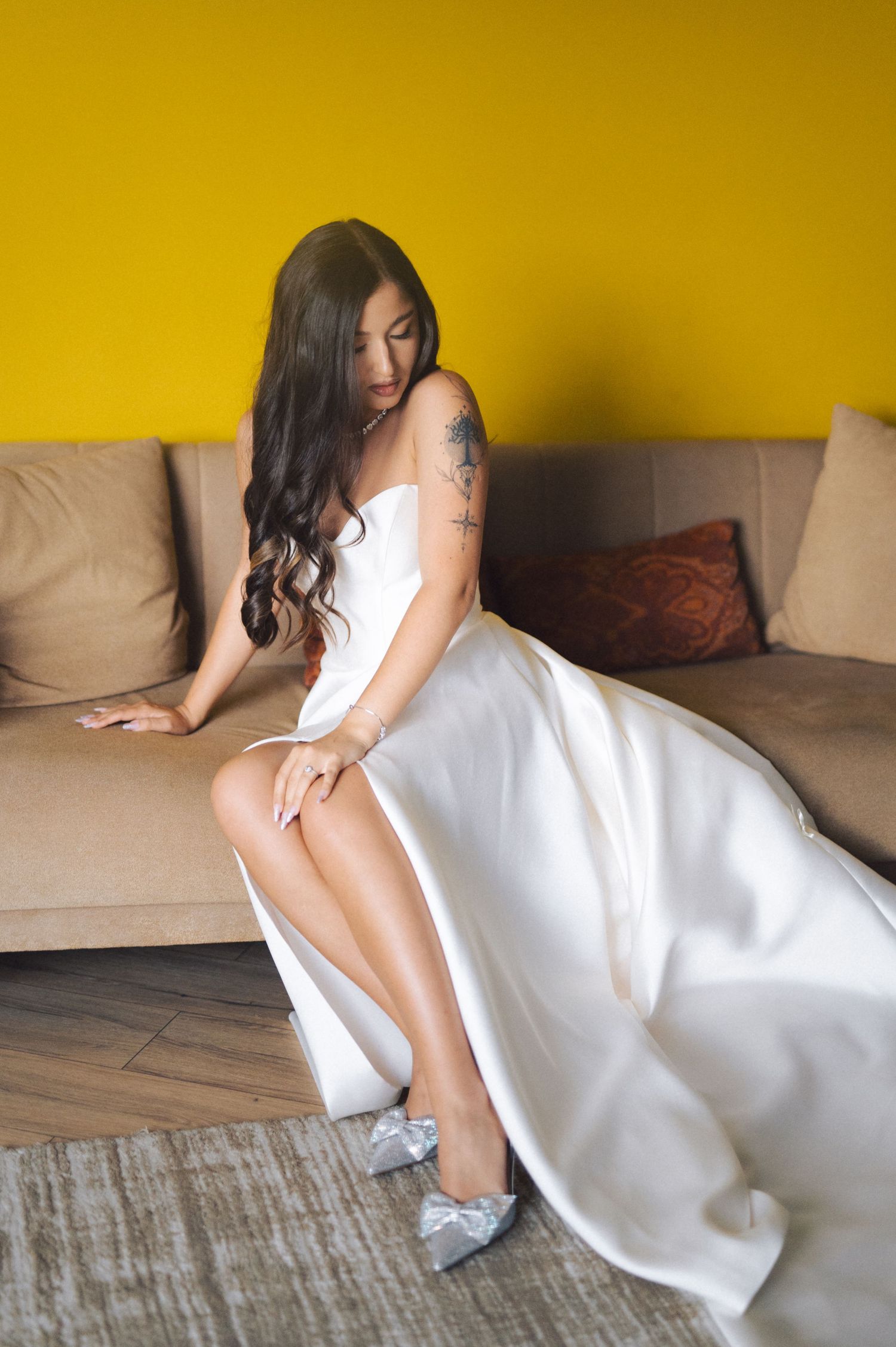Elegant figure in flowing white gown and silver shoes poses on beige couch against vibrant yellow wall.