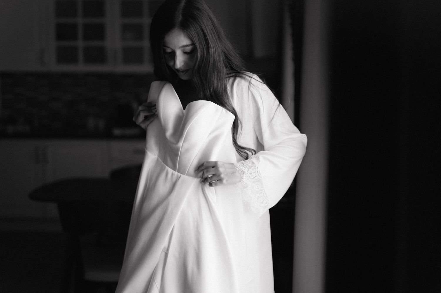 Dramatic black and white portrait of a person wearing a flowy white dress in a dimly lit interior setting.