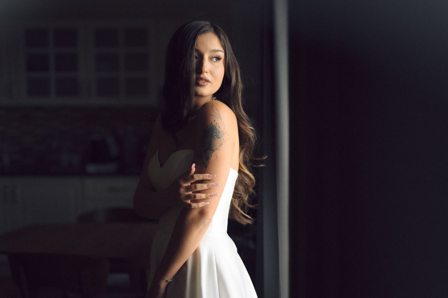 A woman in a white dress poses near a window with dramatic side lighting creating a moody atmosphere.