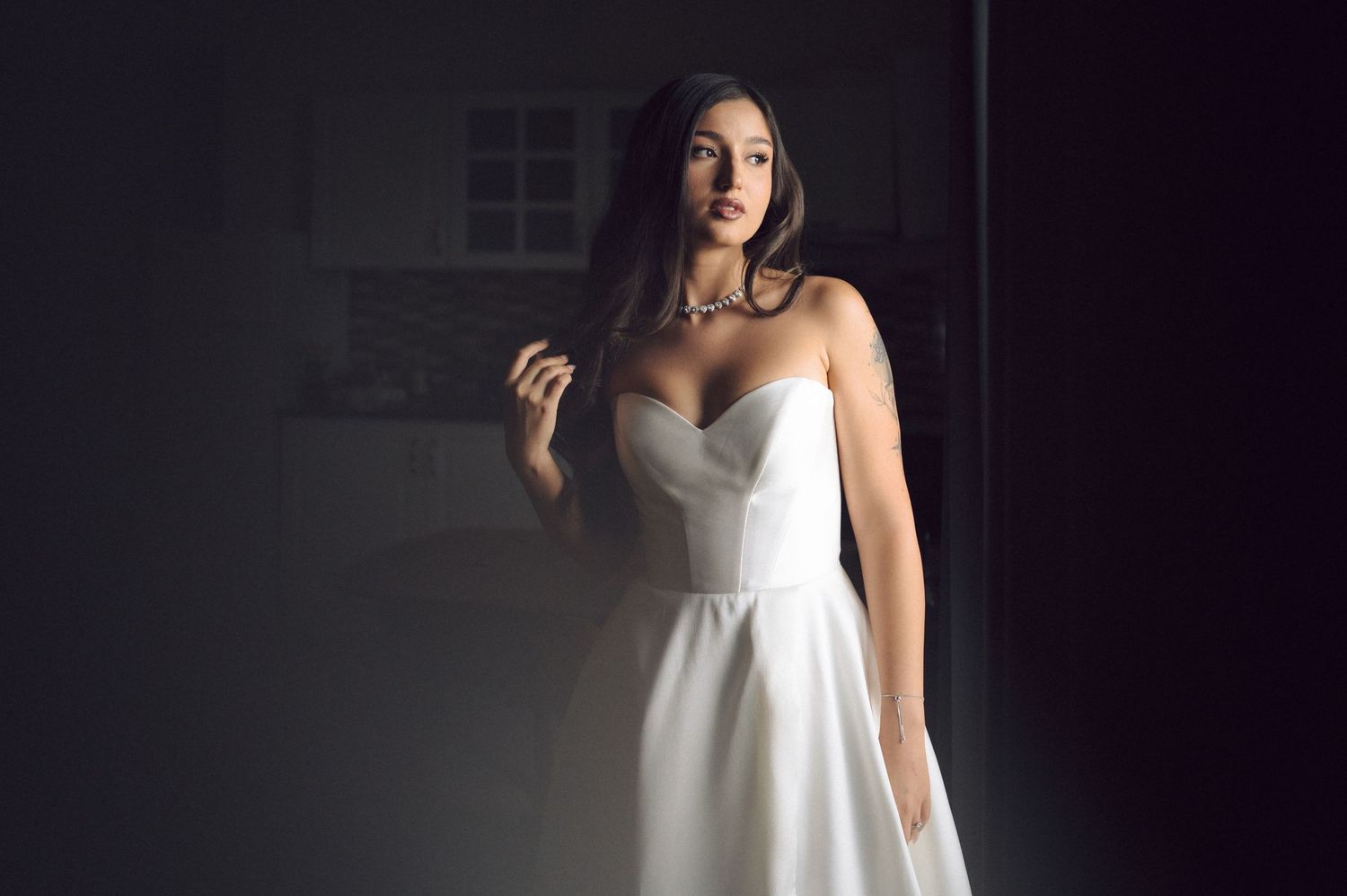 A woman in an elegant white strapless wedding dress poses against a dark background with dramatic lighting.