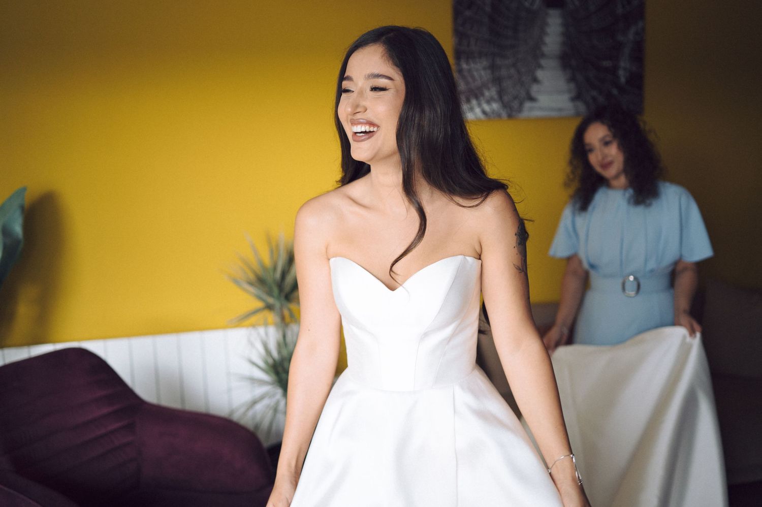 A woman in a white dress sits on a bed during what appears to be a bridal preparation session.