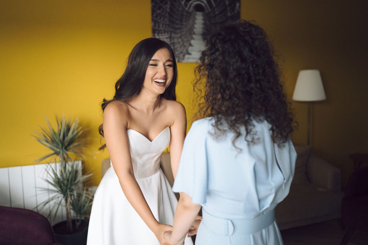 Two people share a joyful moment while preparing in a room with yellow walls and indoor plants.