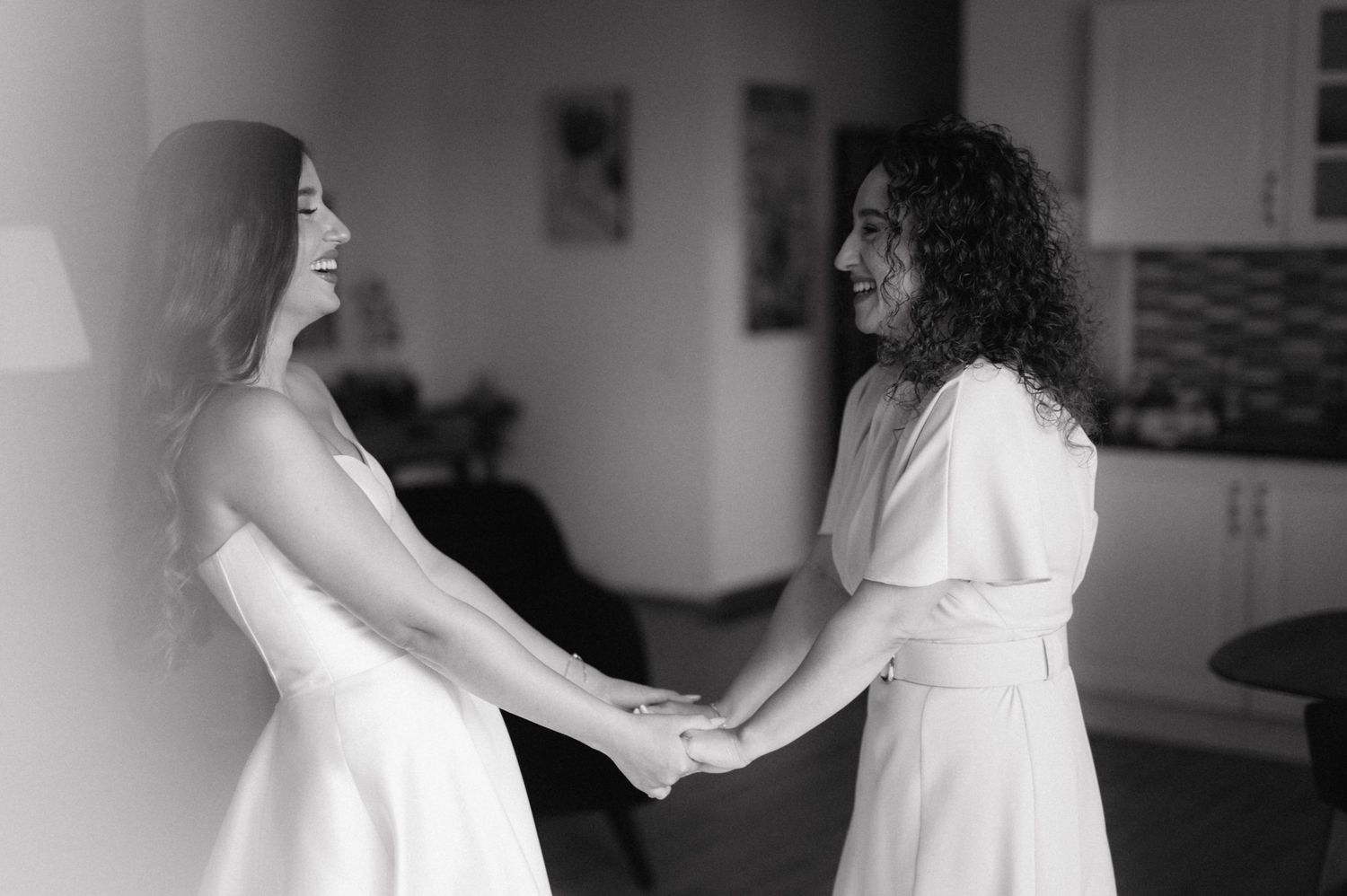 Black and white photo of two people in white dresses holding hands and laughing together indoors.