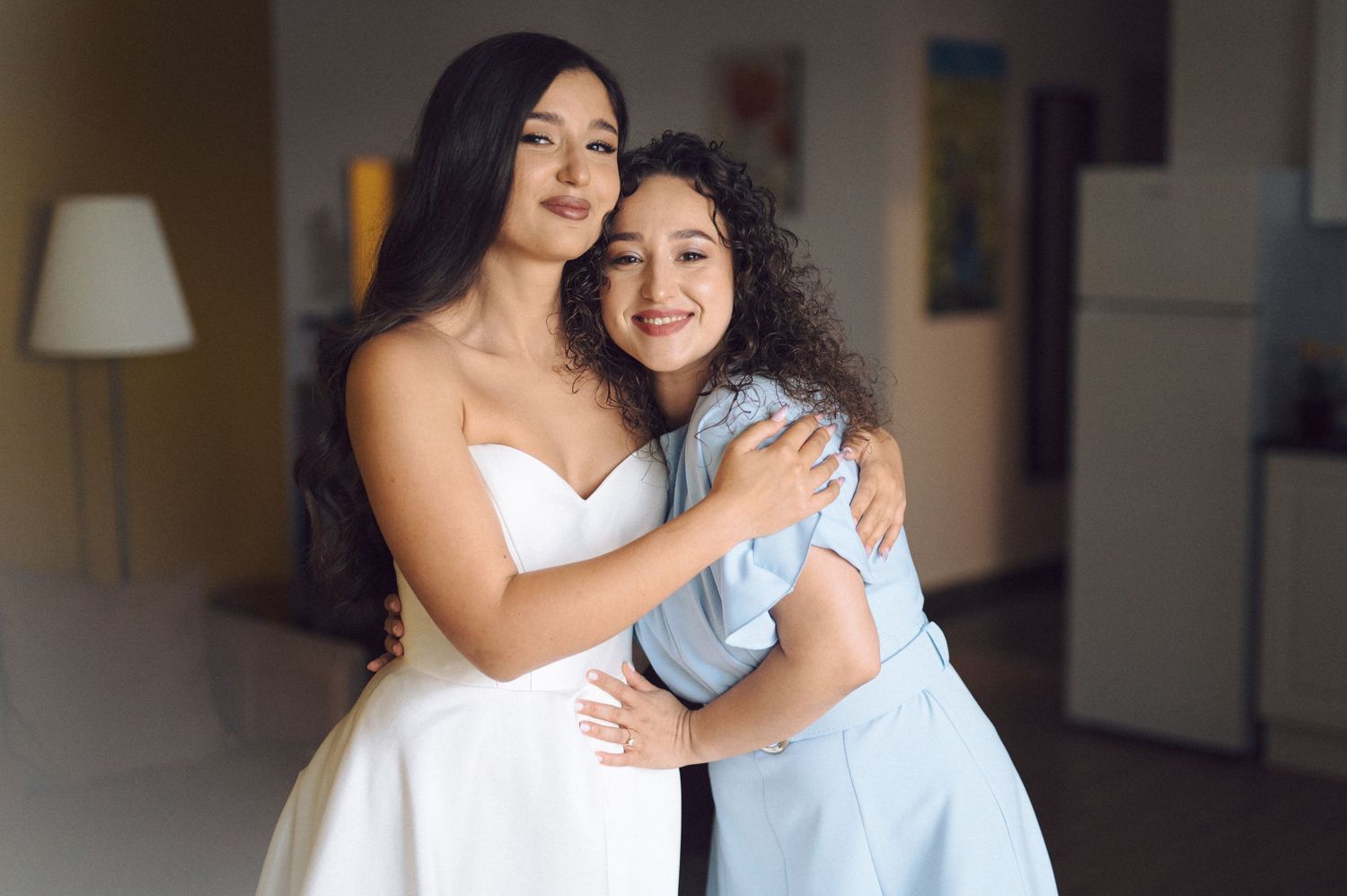Two people share a joyful embrace in formal attire with bright lighting in a hallway setting.