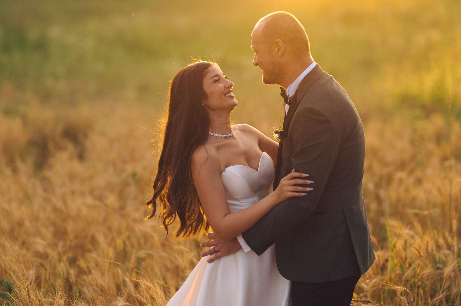 A couple in wedding attire embrace in a golden wheat field at sunset while sharing a romantic moment.