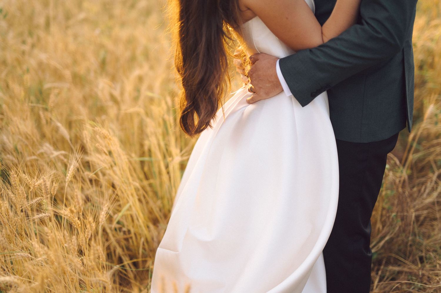 A romantic embrace between a couple in formal attire standing in a golden wheat field at sunset.