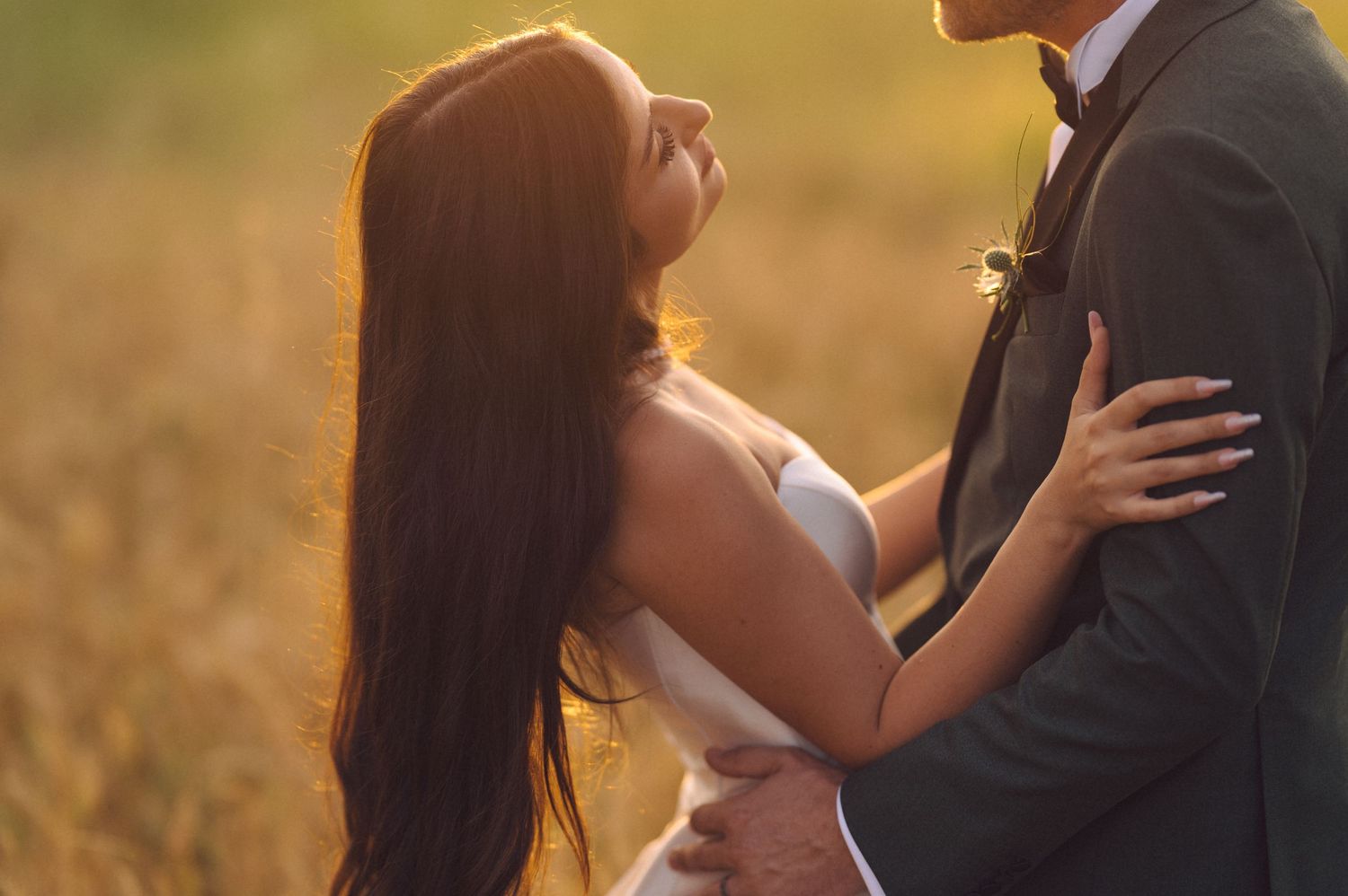 Romantic couple embraces in golden sunlit field during sunset wearing formal wedding attire.