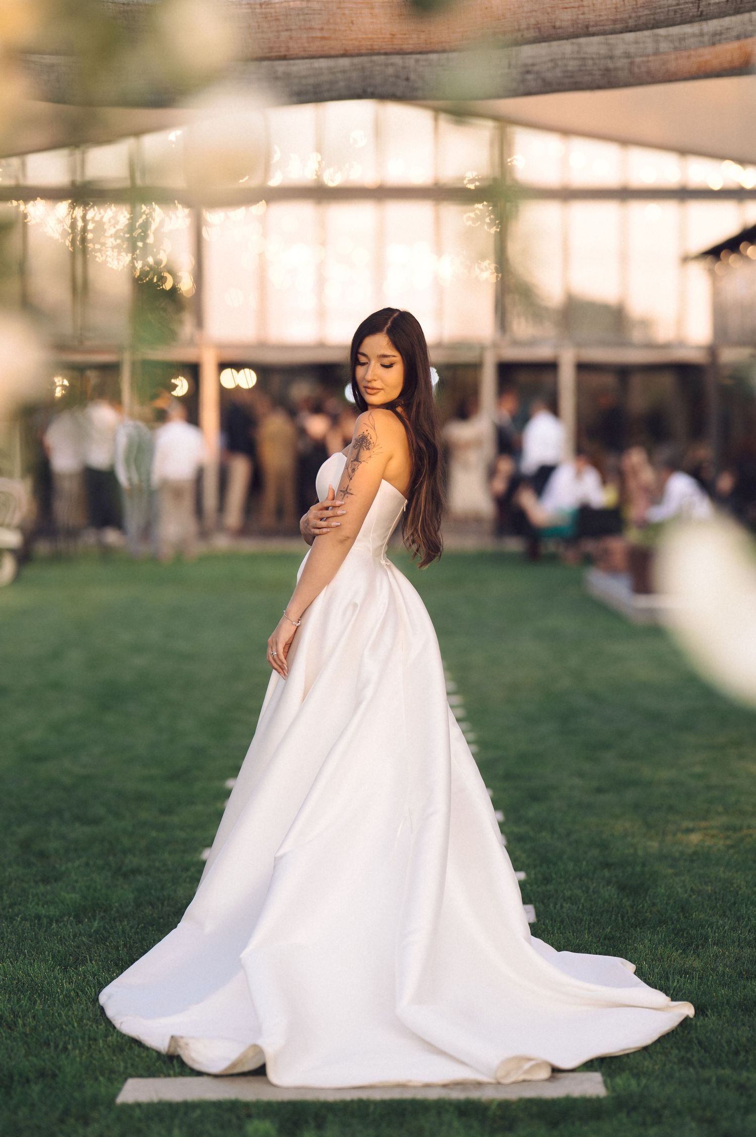A bride in a flowing white wedding gown poses elegantly on a lawn with guests visible in the background.