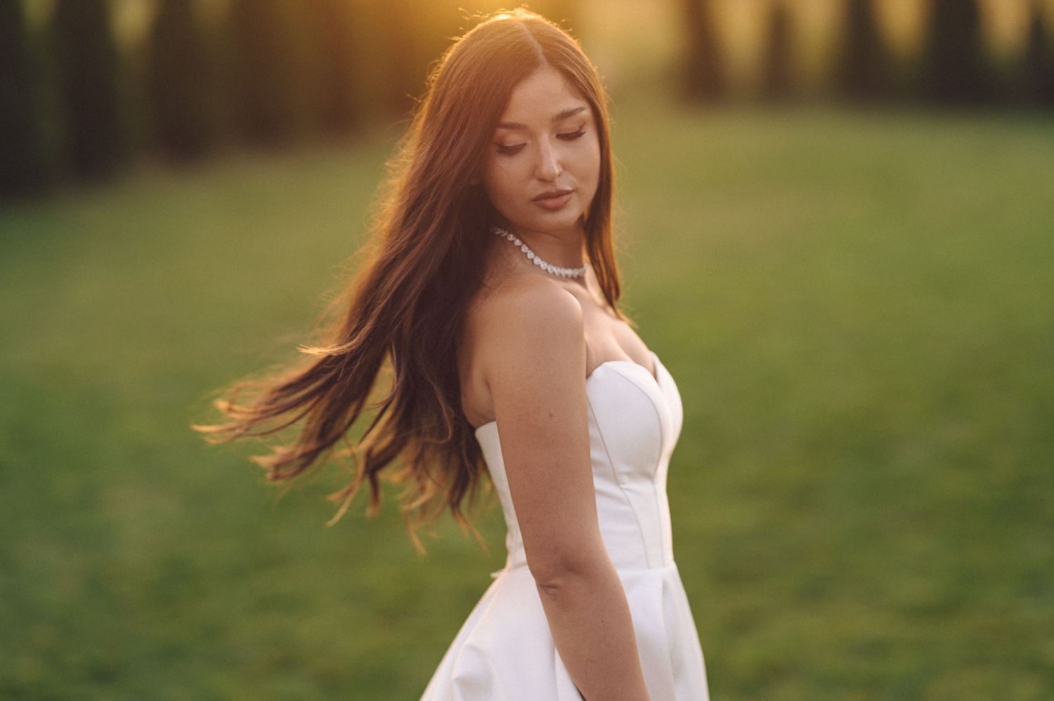 A woman in a flowing white dress stands in a sunlit grassy field with her hair flowing in the breeze during golden hour.