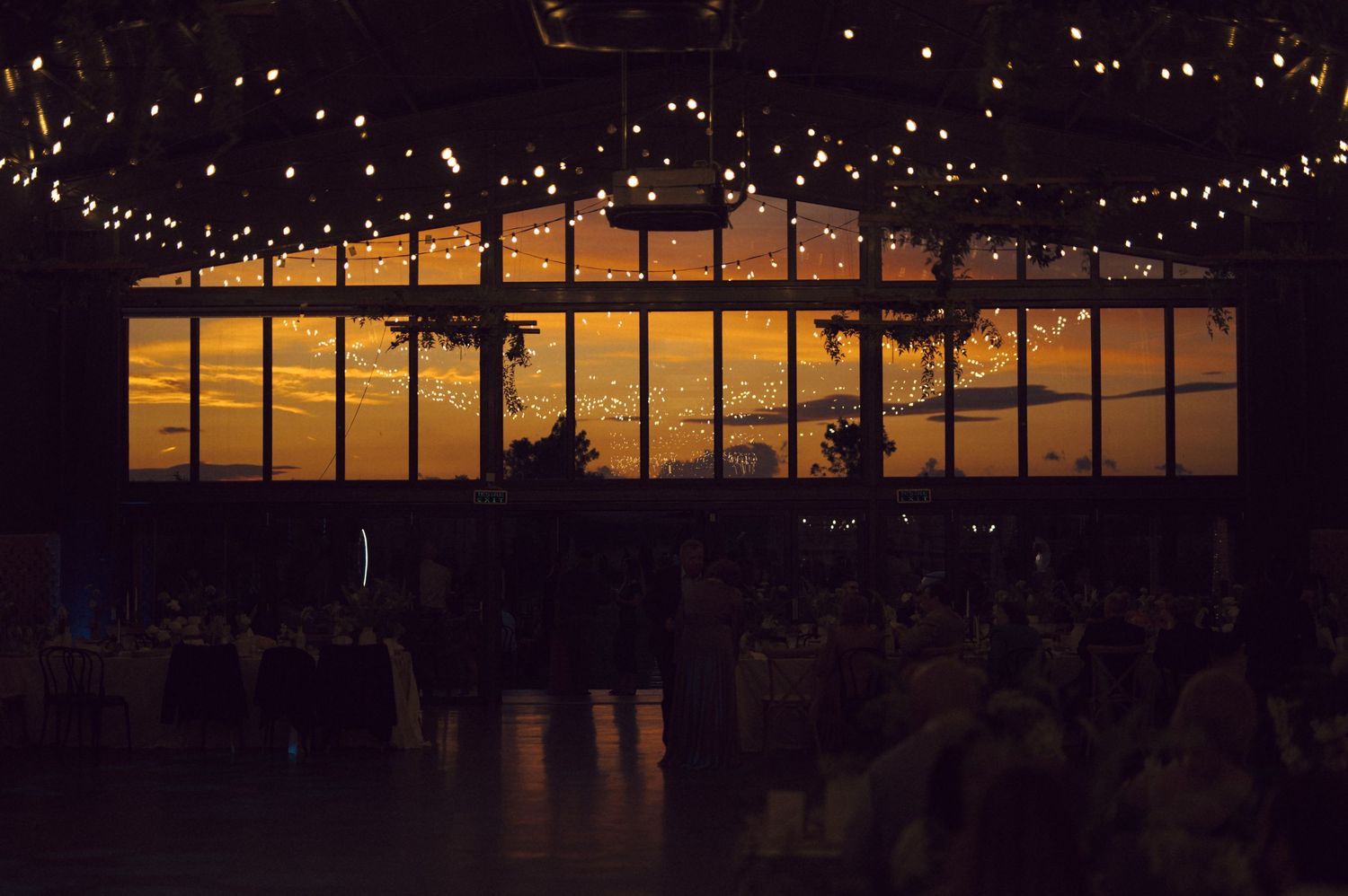 Illuminated glass windows at dusk create a warm glow beneath string lights at an evening event venue.