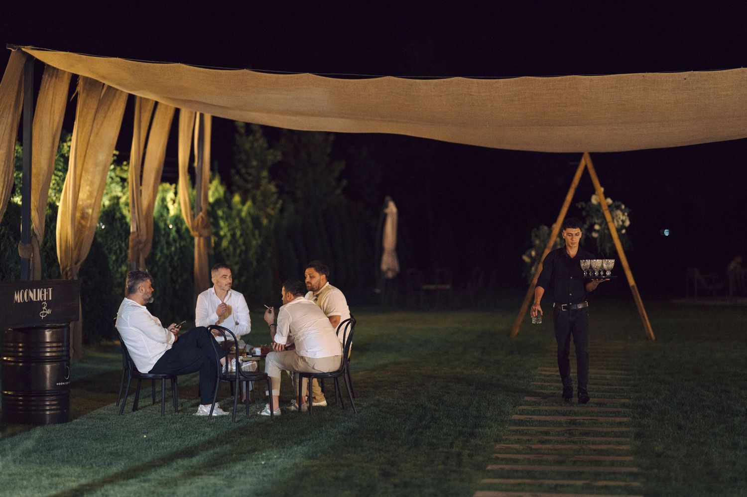 Small group gathered under outdoor tent canopy at night with ambient lighting and lawn seating.