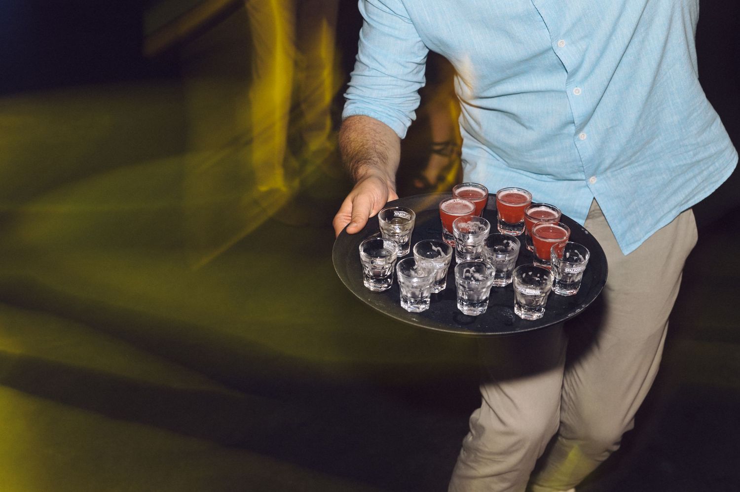 Server carrying tray of shot glasses at dimly lit event.