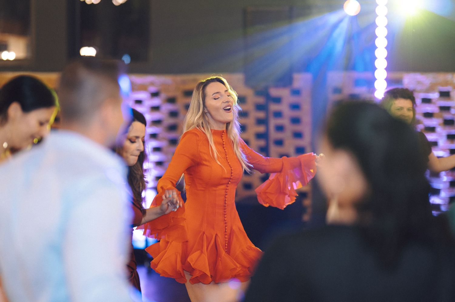Woman in orange dress dancing energetically at party with colorful lighting.