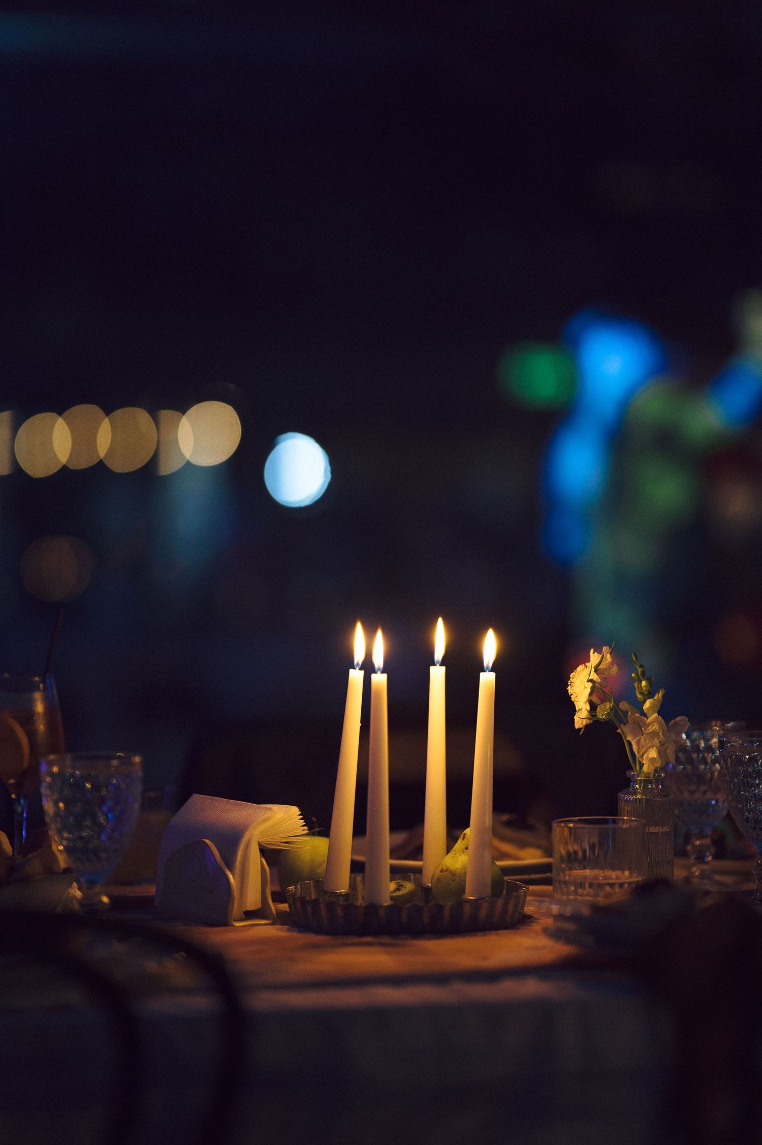 Four white candles burning on an elegantly set dinner table with bokeh lights in the background.