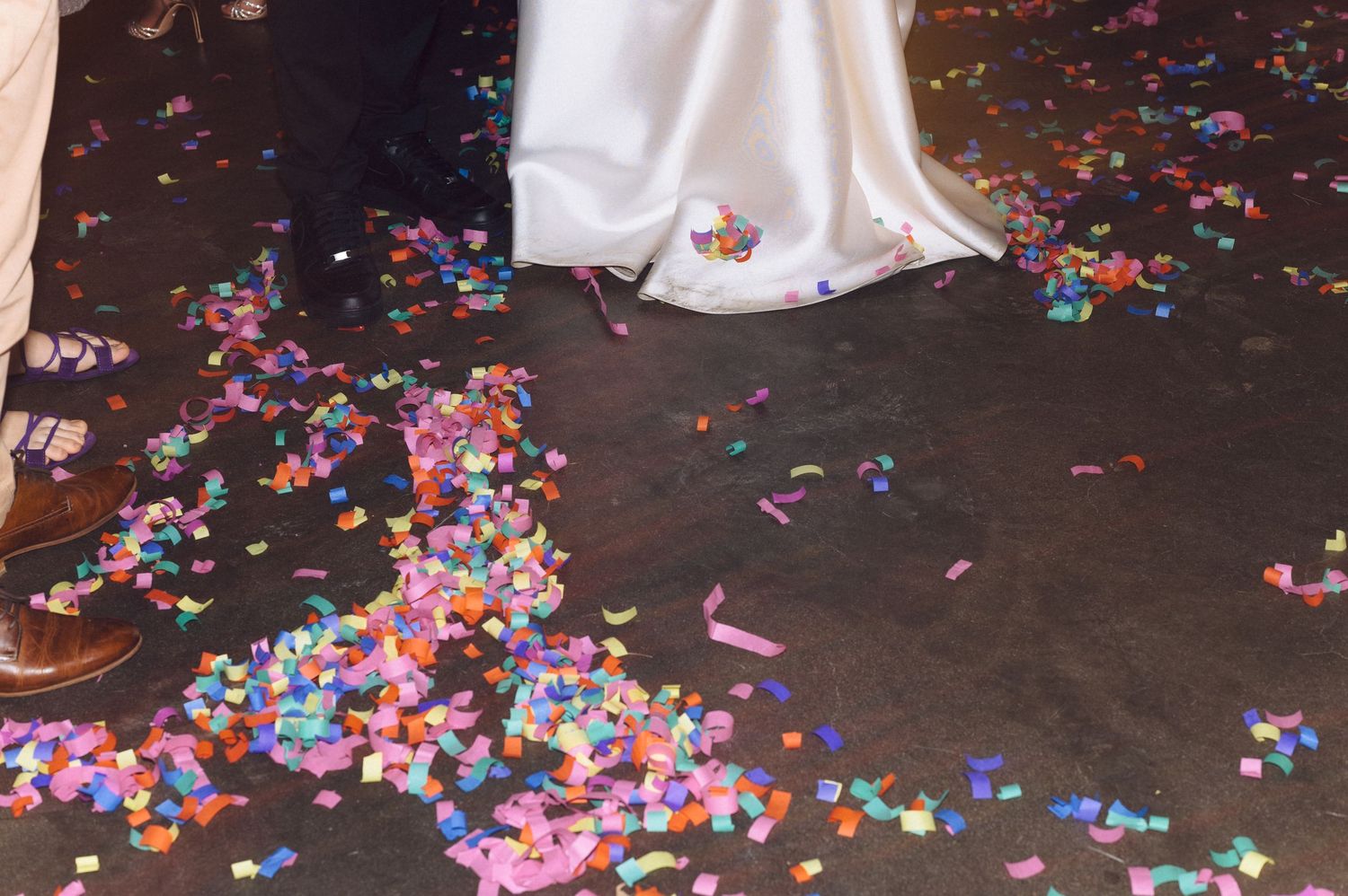 The hem of a white wedding dress surrounded by scattered colorful confetti on a dark floor.
