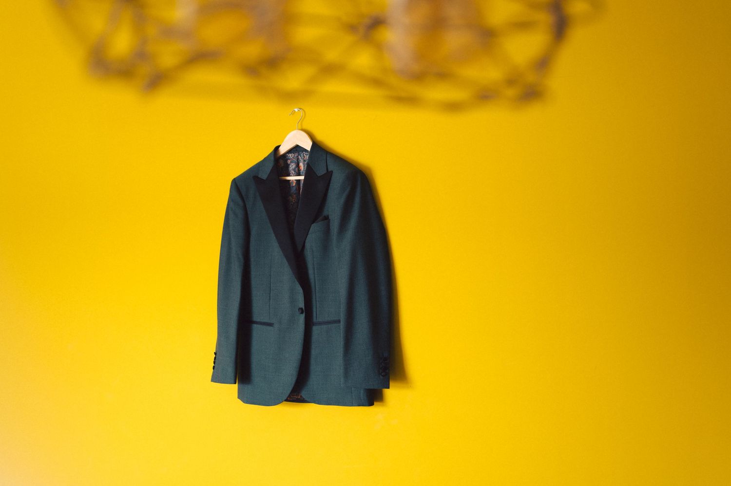 Dark green blazer jacket hanging on white hanger against vibrant yellow wall with decorative element above.