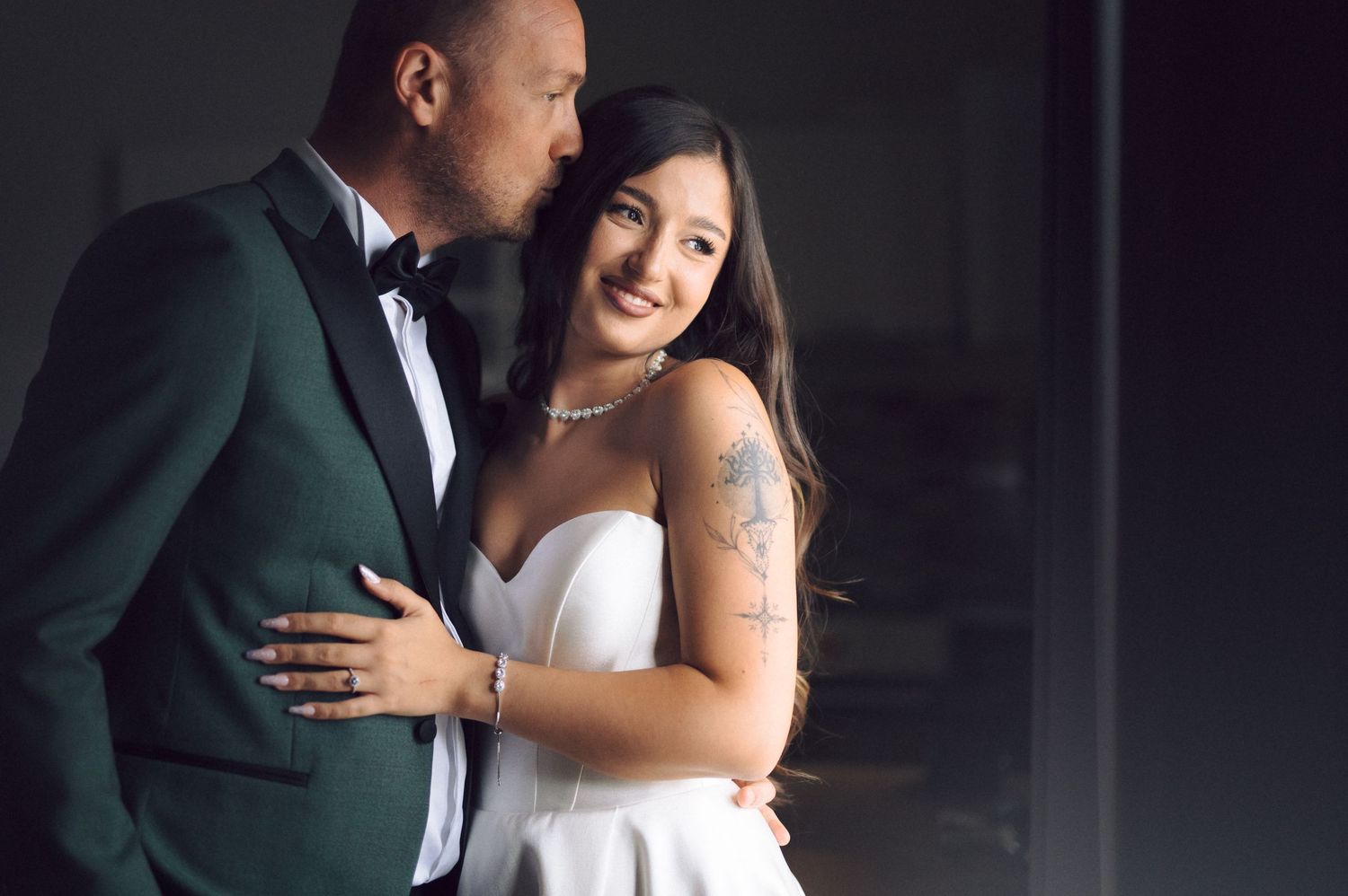 A romantic couple embraces in formal wedding attire, with the groom in a dark green tuxedo and bride in a white dress.