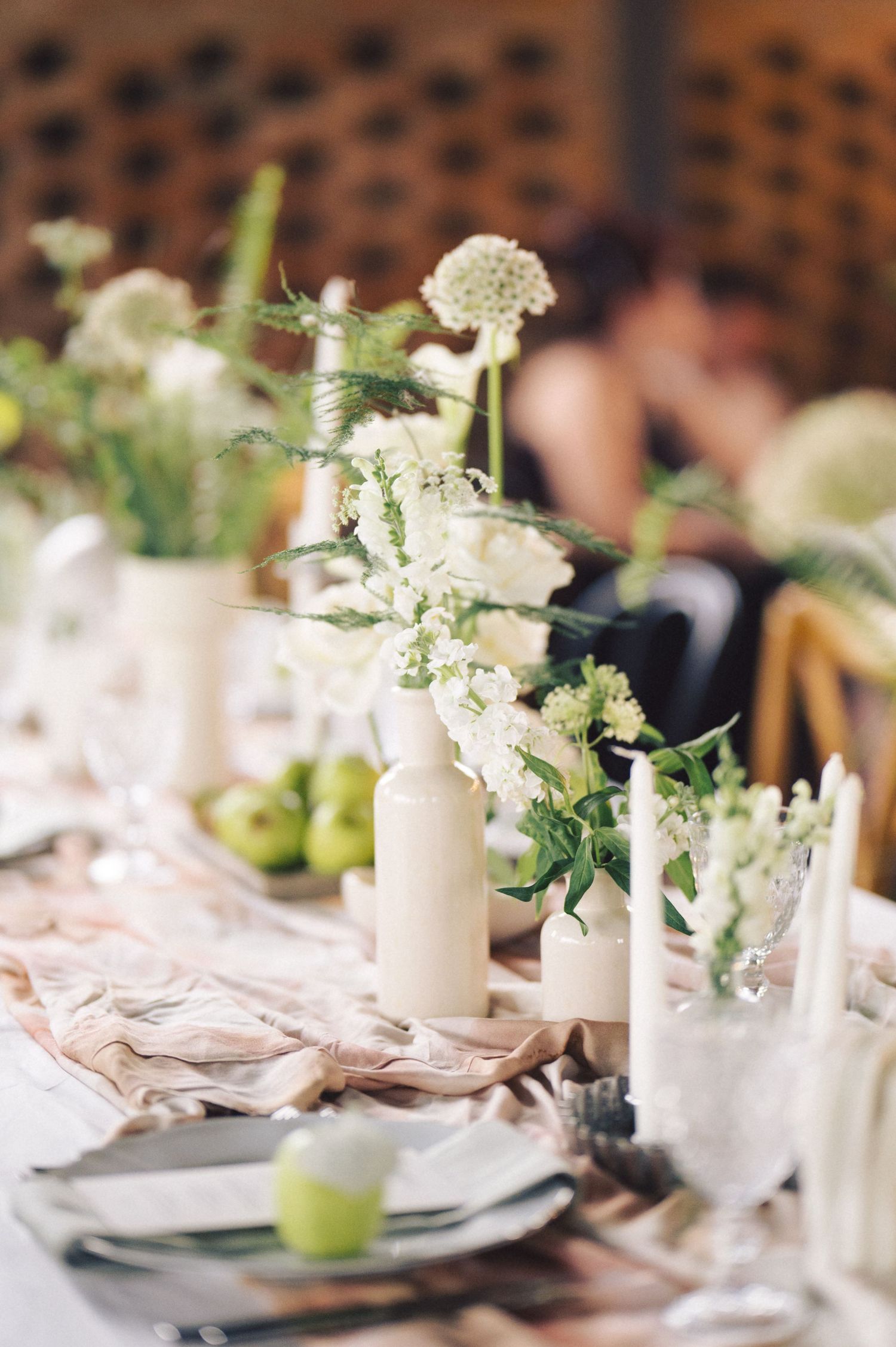 White floral centerpieces in delicate vases adorn an elegant wedding reception table with clean place settings and linens.