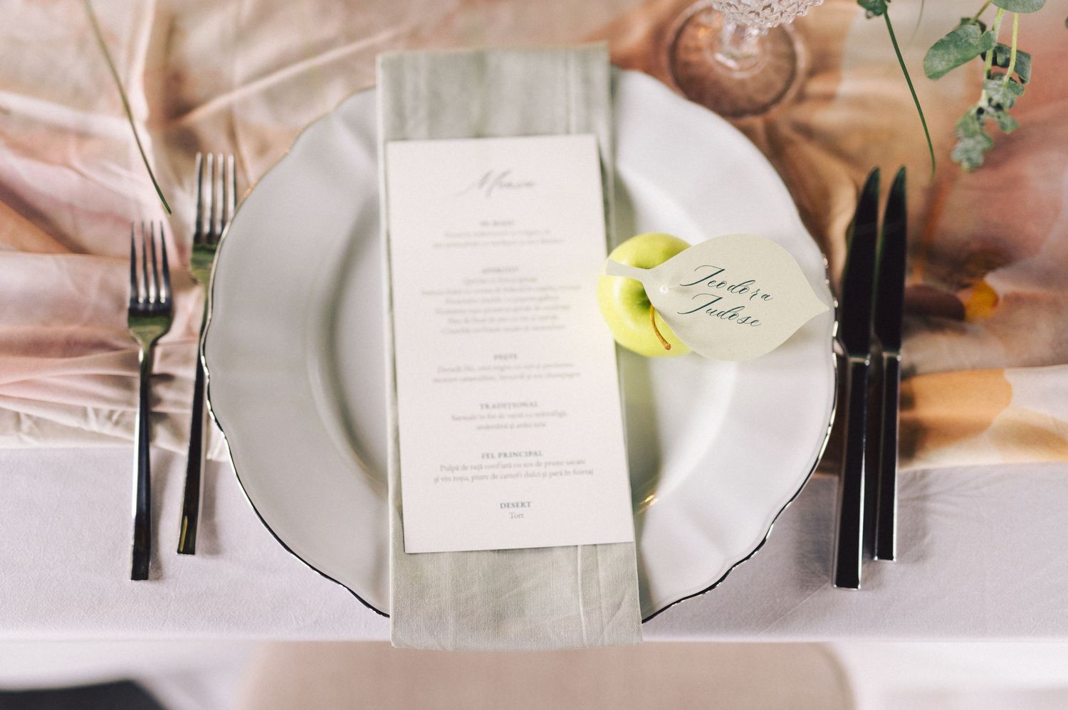 Elegant place setting with white plate, menu card, and black silverware on a blush pink tablecloth.
