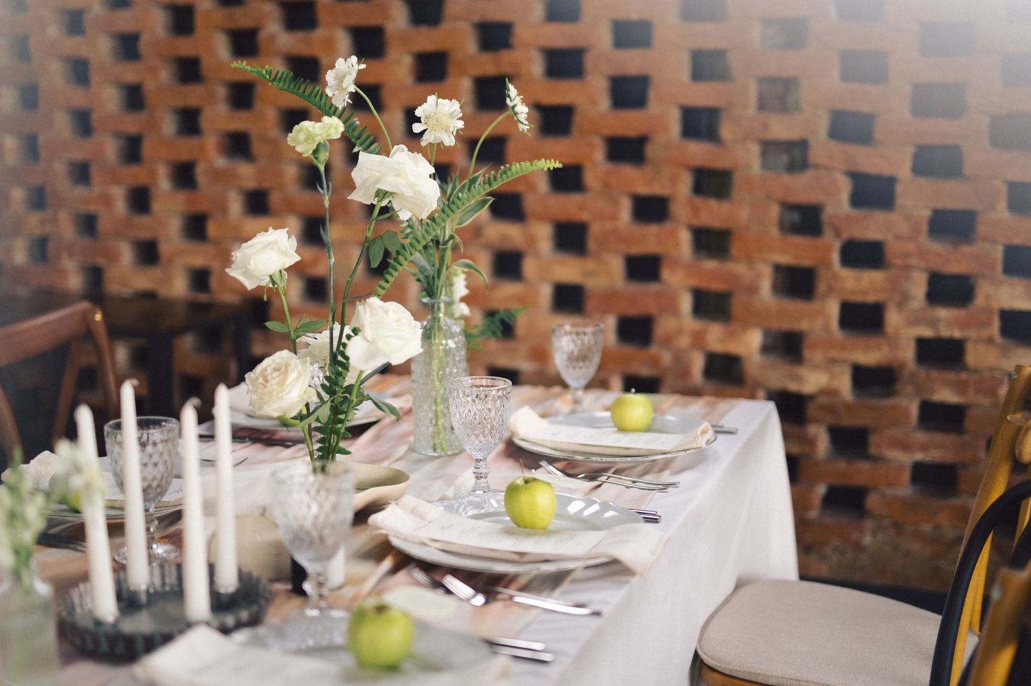 An elegant table setting with white flowers in a glass vase, candles, and green accents against a brick wall background.