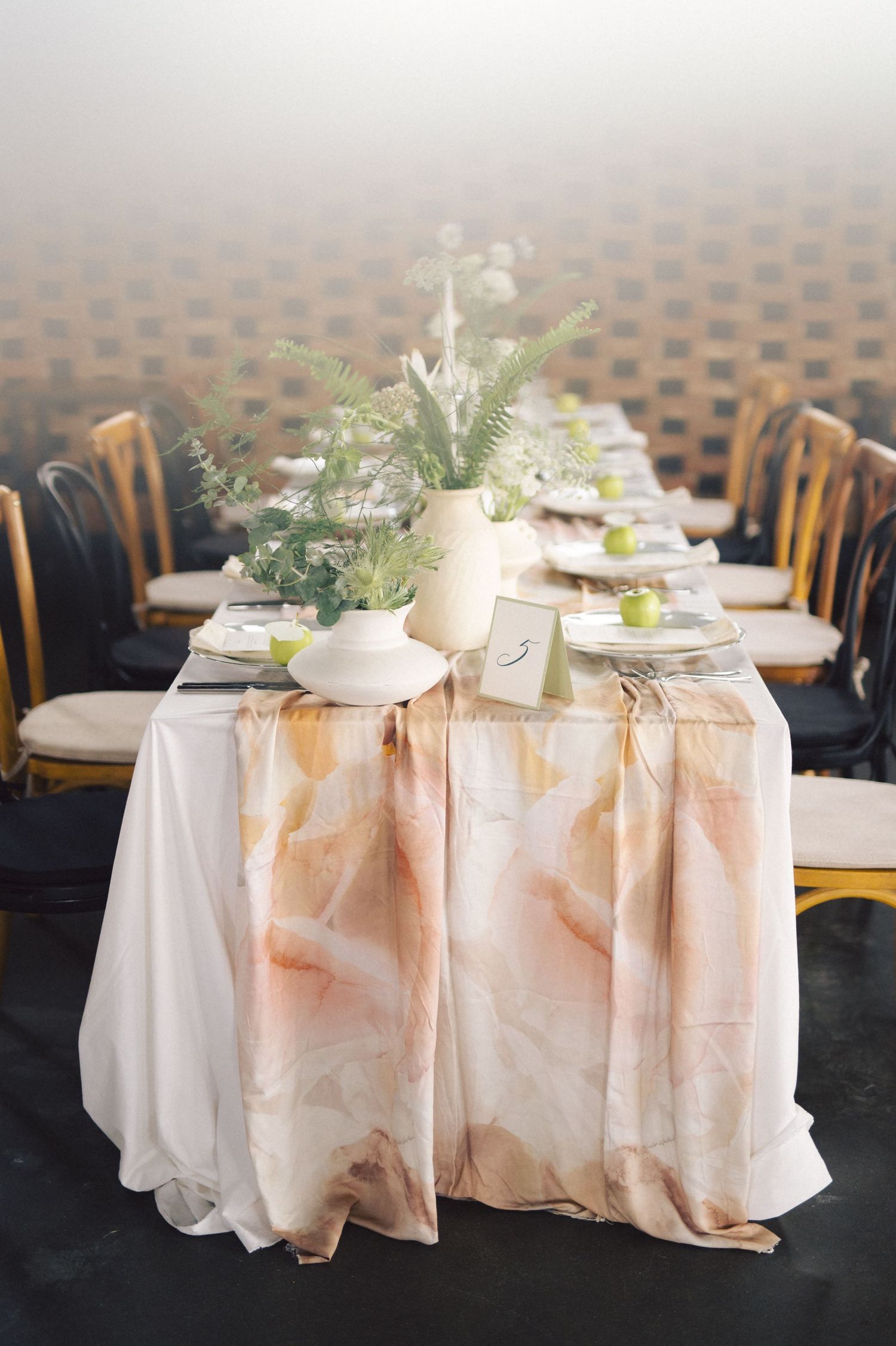 An elegant table setting with a peach ombre runner, delicate greenery centerpieces, and bistro chairs at a formal event.