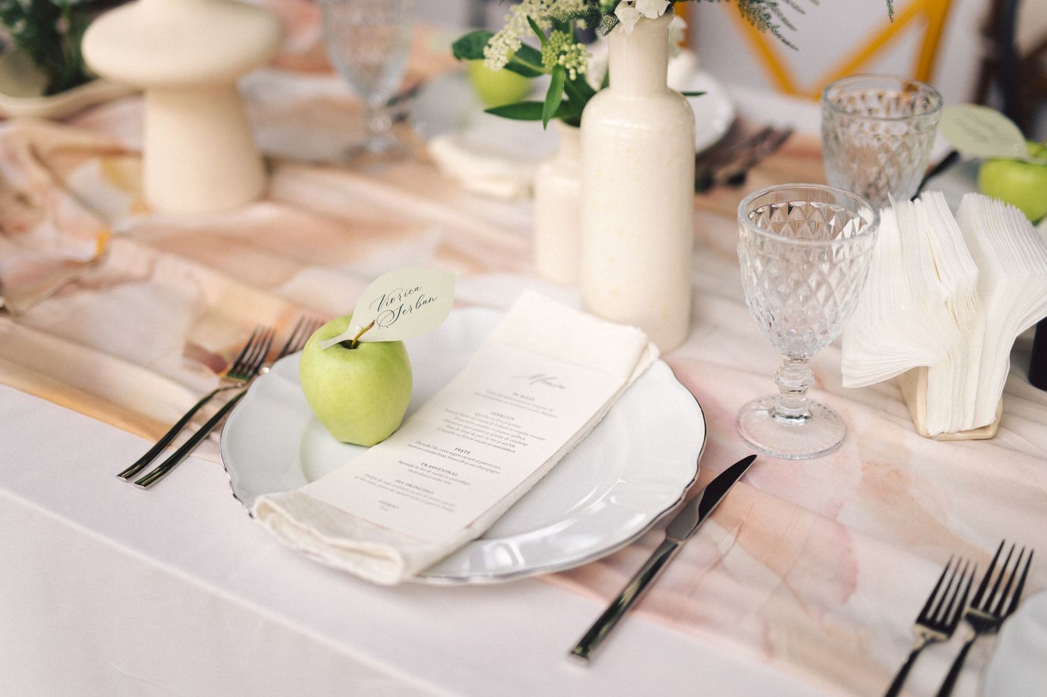 An elegant place setting with white plates, crystal glassware, and a green apple on a pale pink tablecloth.