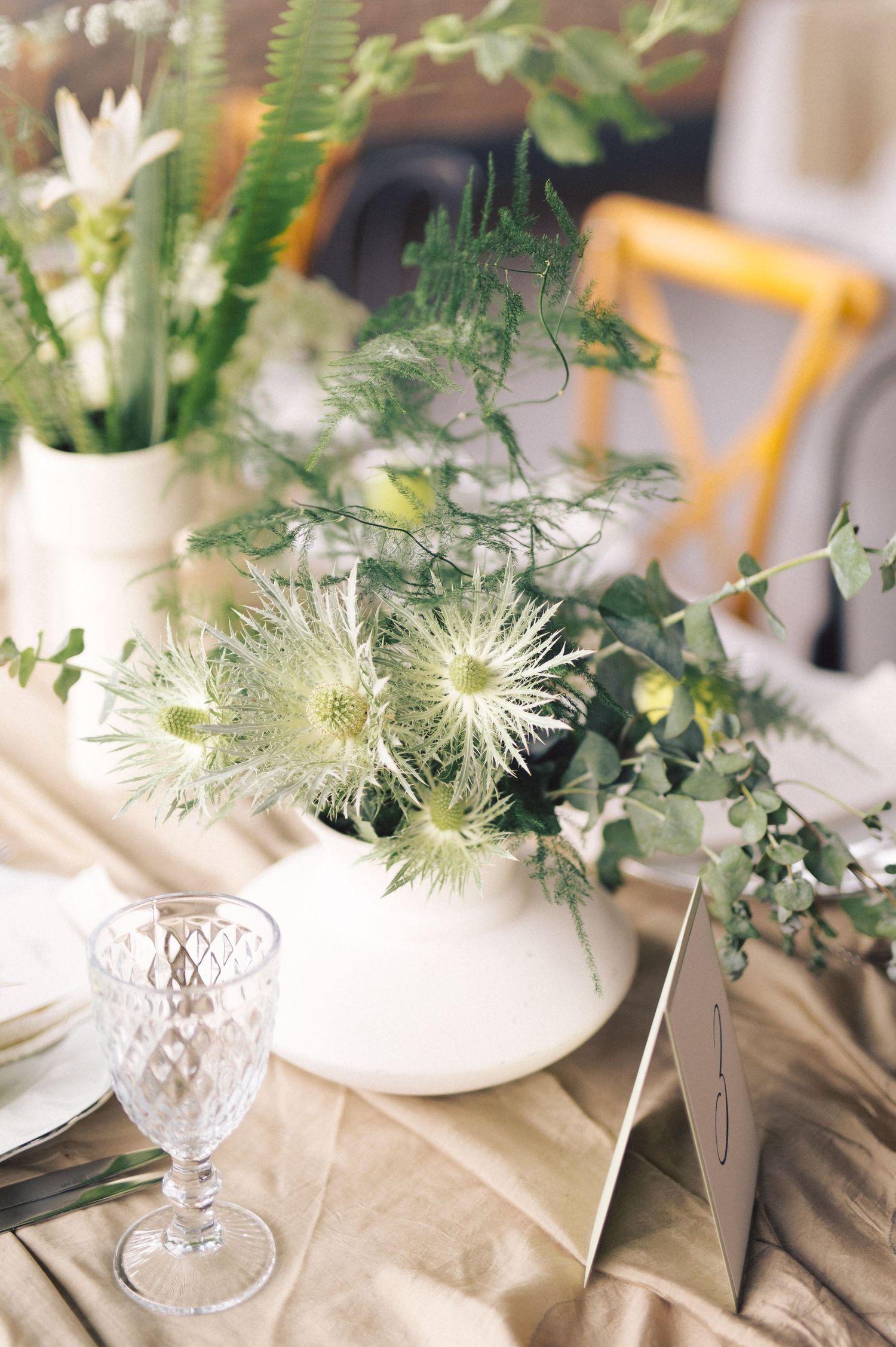 Elegant table setting with delicate white floral centerpieces, ferns, and crystal glassware on a neutral linen tablecloth.