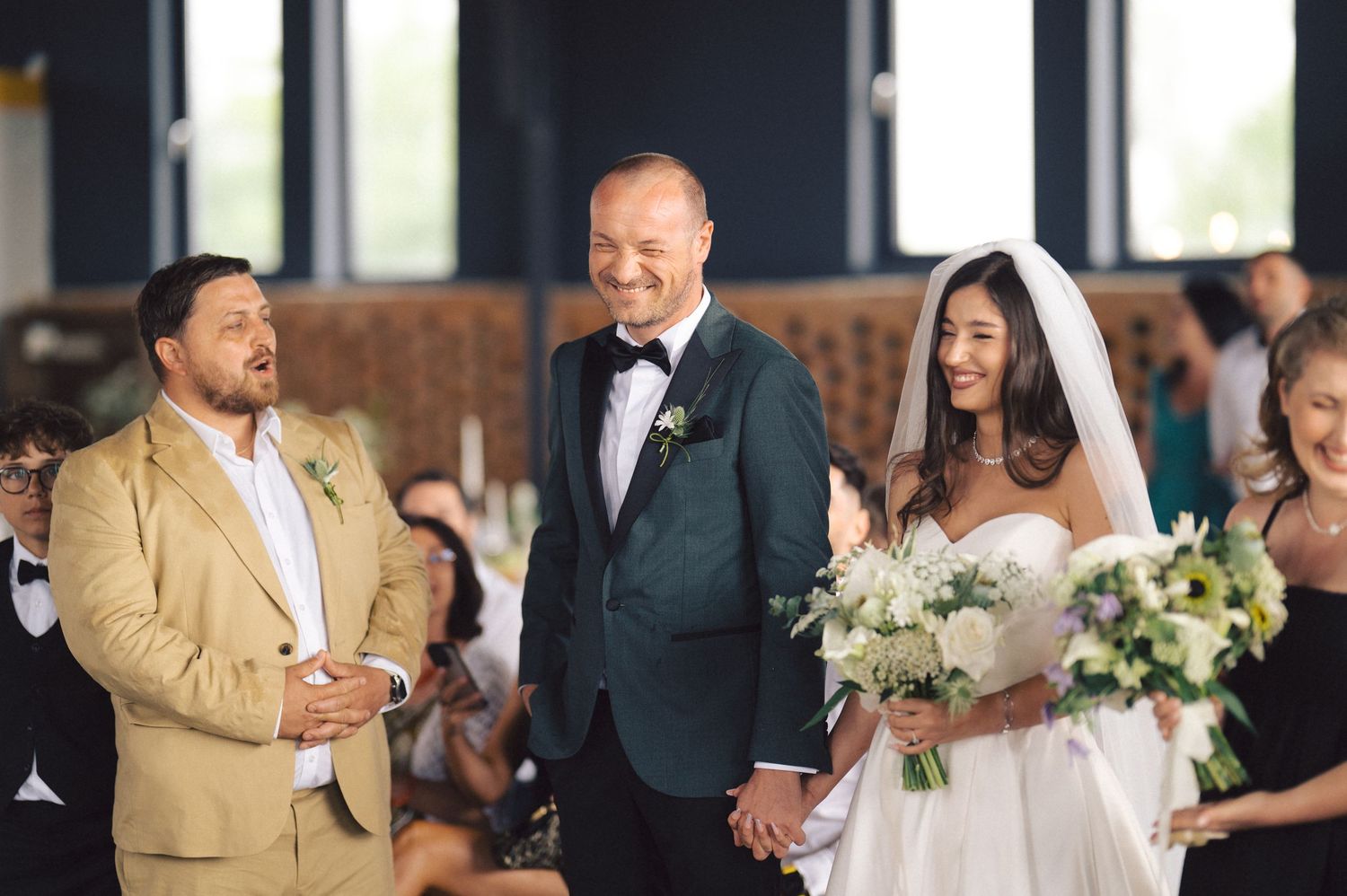 A joyful wedding ceremony inside a venue with elegant white floral bouquets and guests in formal attire.