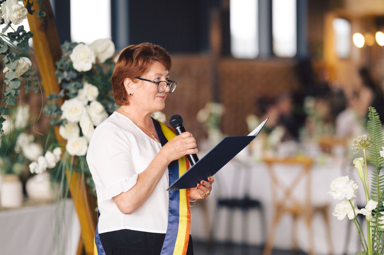 Person in a white shirt wearing glasses speaks from a podium at an elegant event decorated with white floral arrangements.