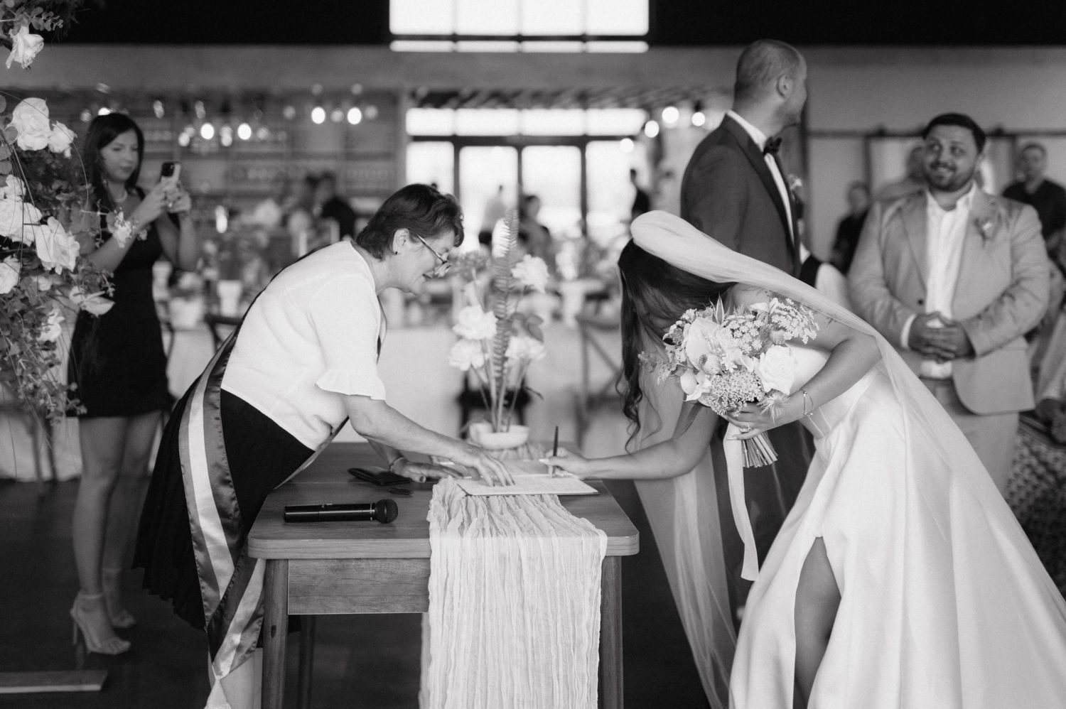 A bride bends down at a rustic wooden table during what appears to be a wedding reception moment captured in black and white.
