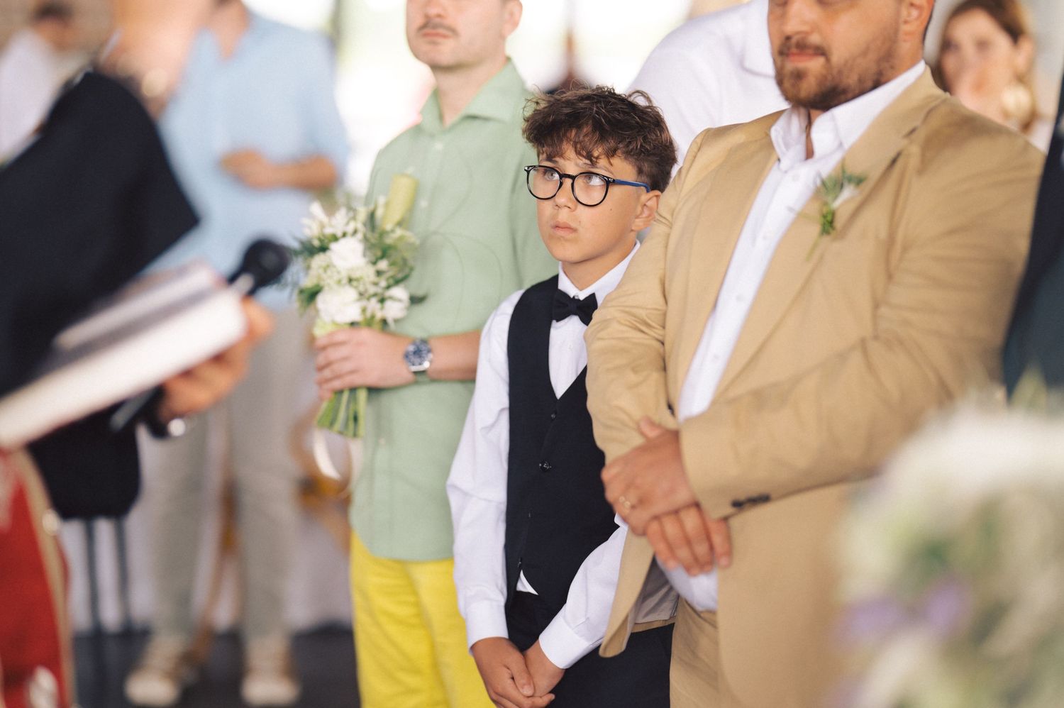 A young ring bearer in a black vest and bowtie stands attentively among wedding party members during a ceremony.