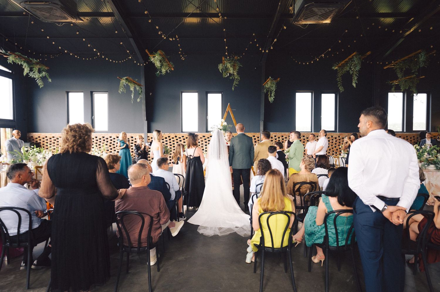 Indoor wedding ceremony in a rustic venue with string lights and hanging greenery above guests seated in rows.