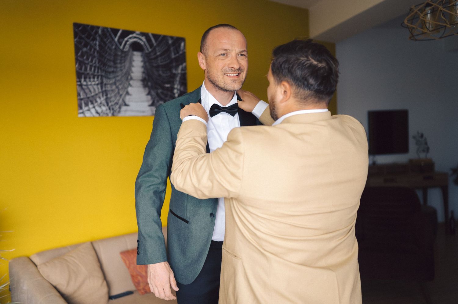 Someone helps adjust a bowtie on a person wearing a formal sage green suit jacket against a yellow wall.
