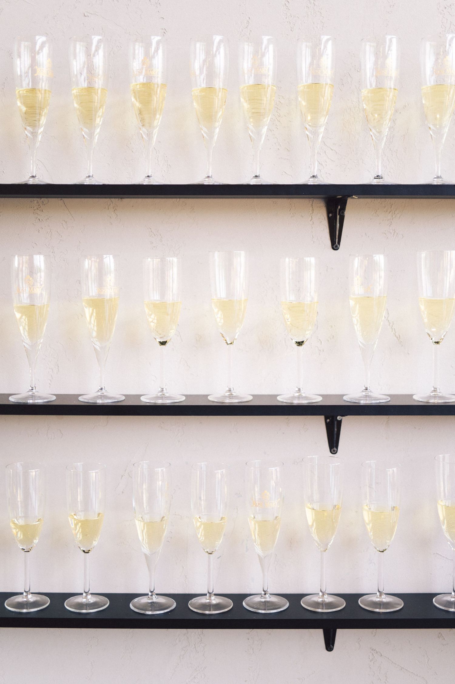 Rows of elegant champagne flutes and wine glasses arranged on black shelving against a white wall.