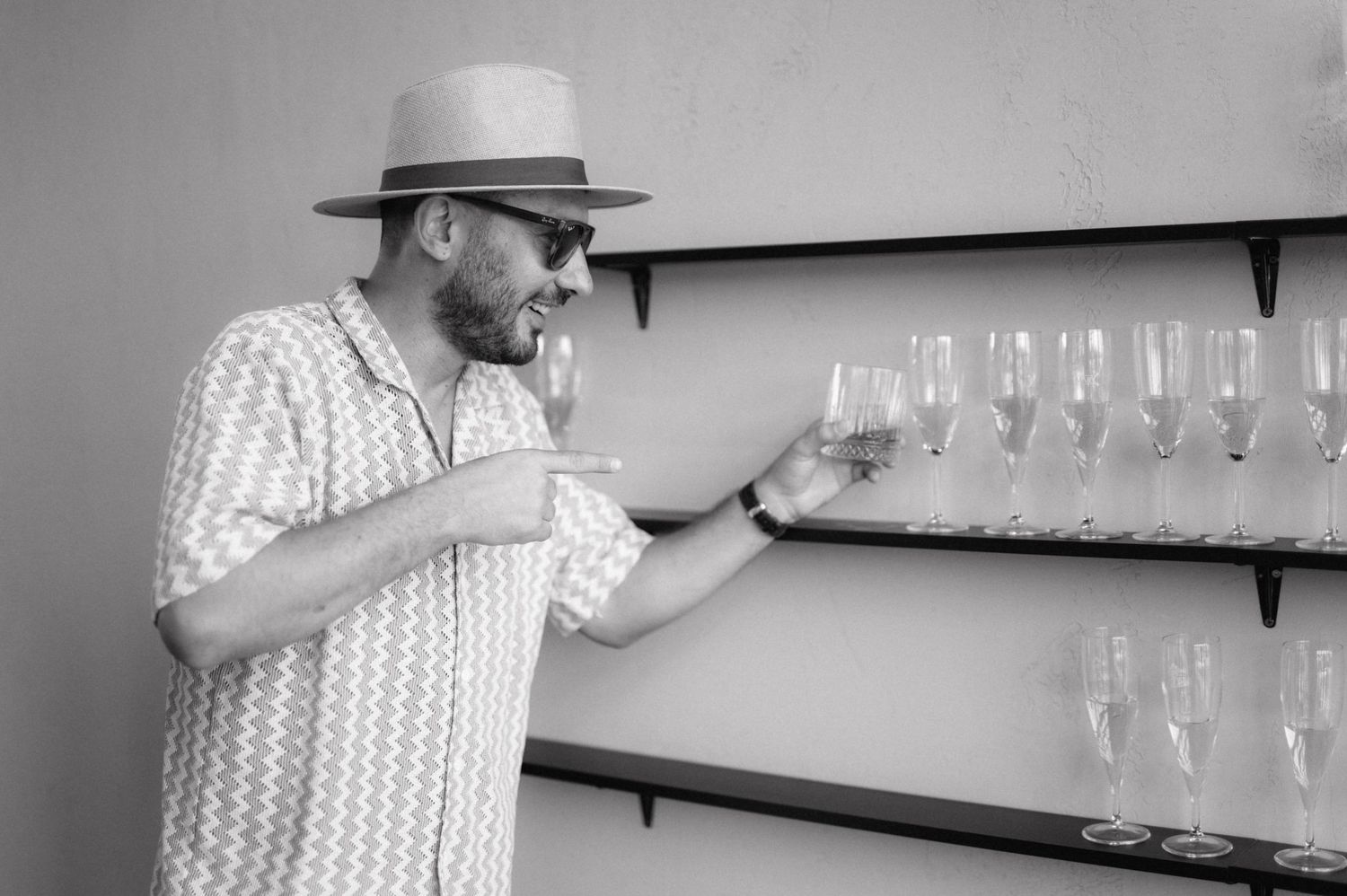 A person wearing a plaid shirt and hat examines wine glasses displayed on wall-mounted shelves.