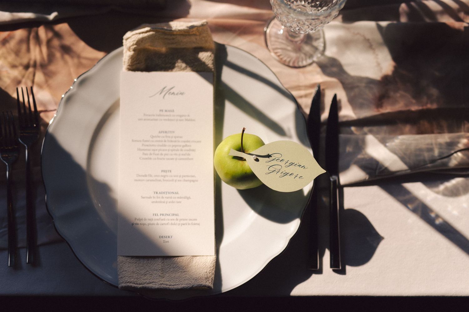 Wedding menu cards set on elegant table settings with sunlight casting shadows across white linens and crystal glassware.