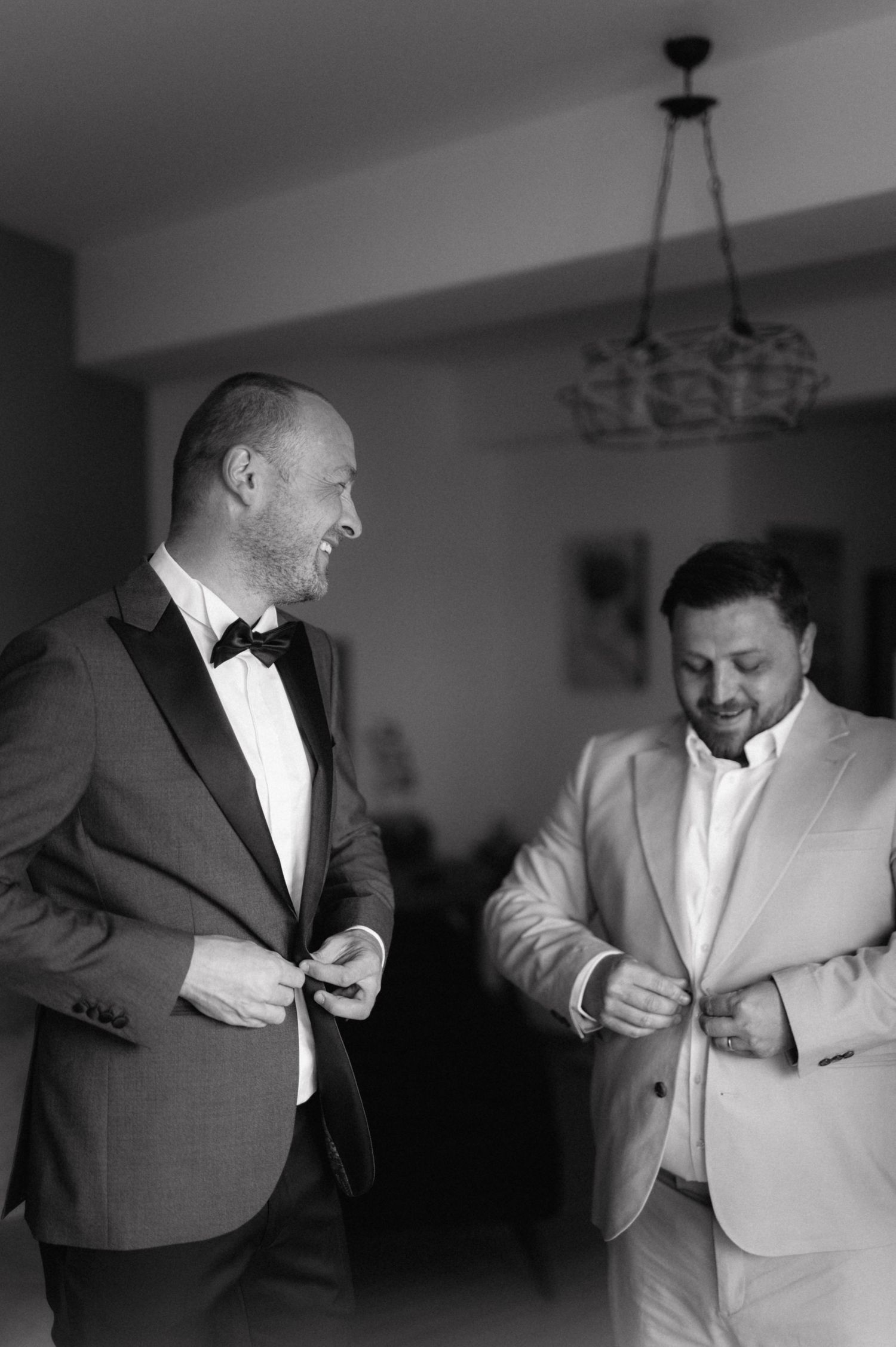Two men in formal attire share a lighthearted moment while getting dressed, one in dark suit and one in light suit.