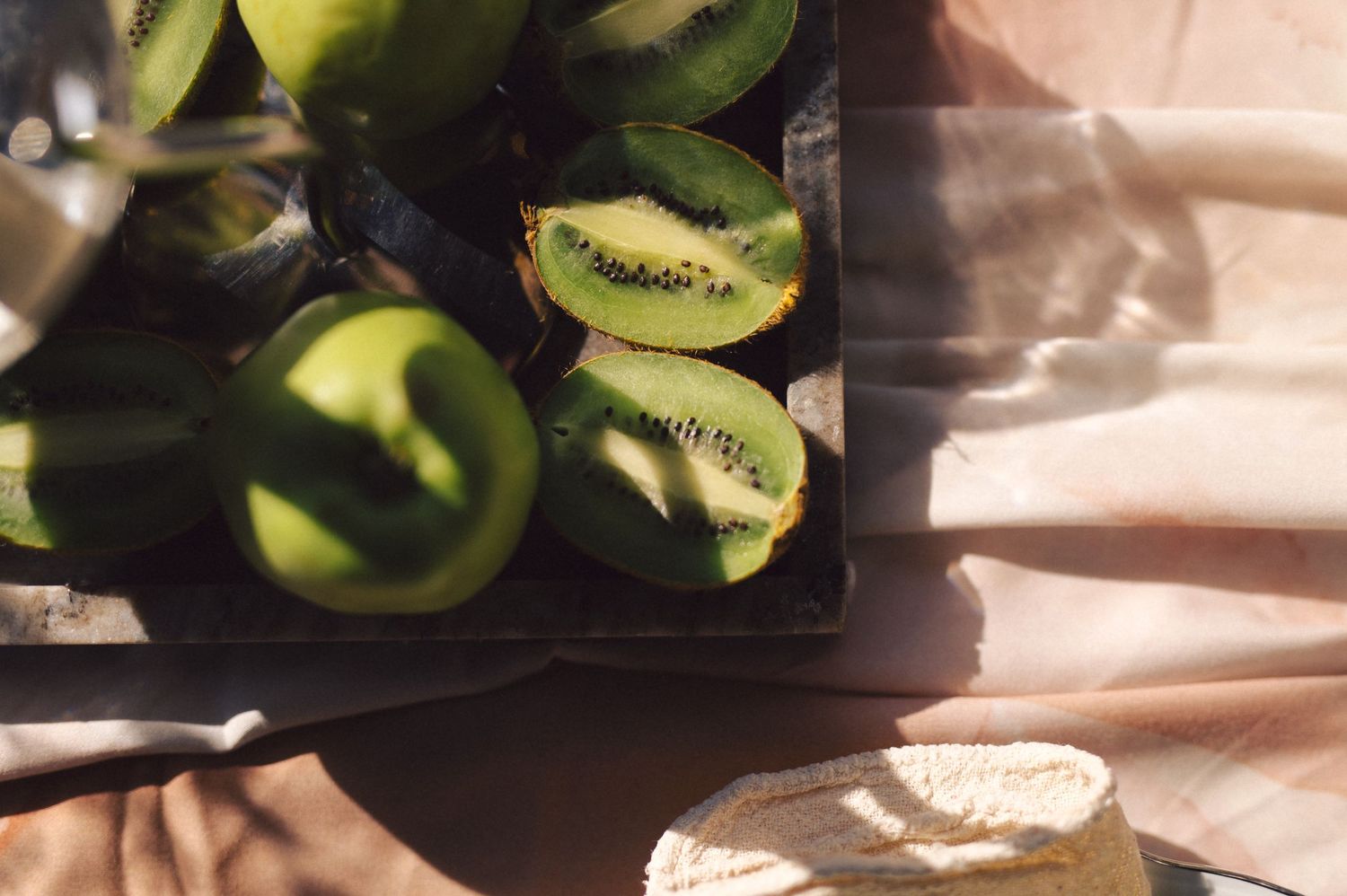 Fresh sliced kiwis and other green fruits arranged on a dark surface with soft pink fabric in sunlight.