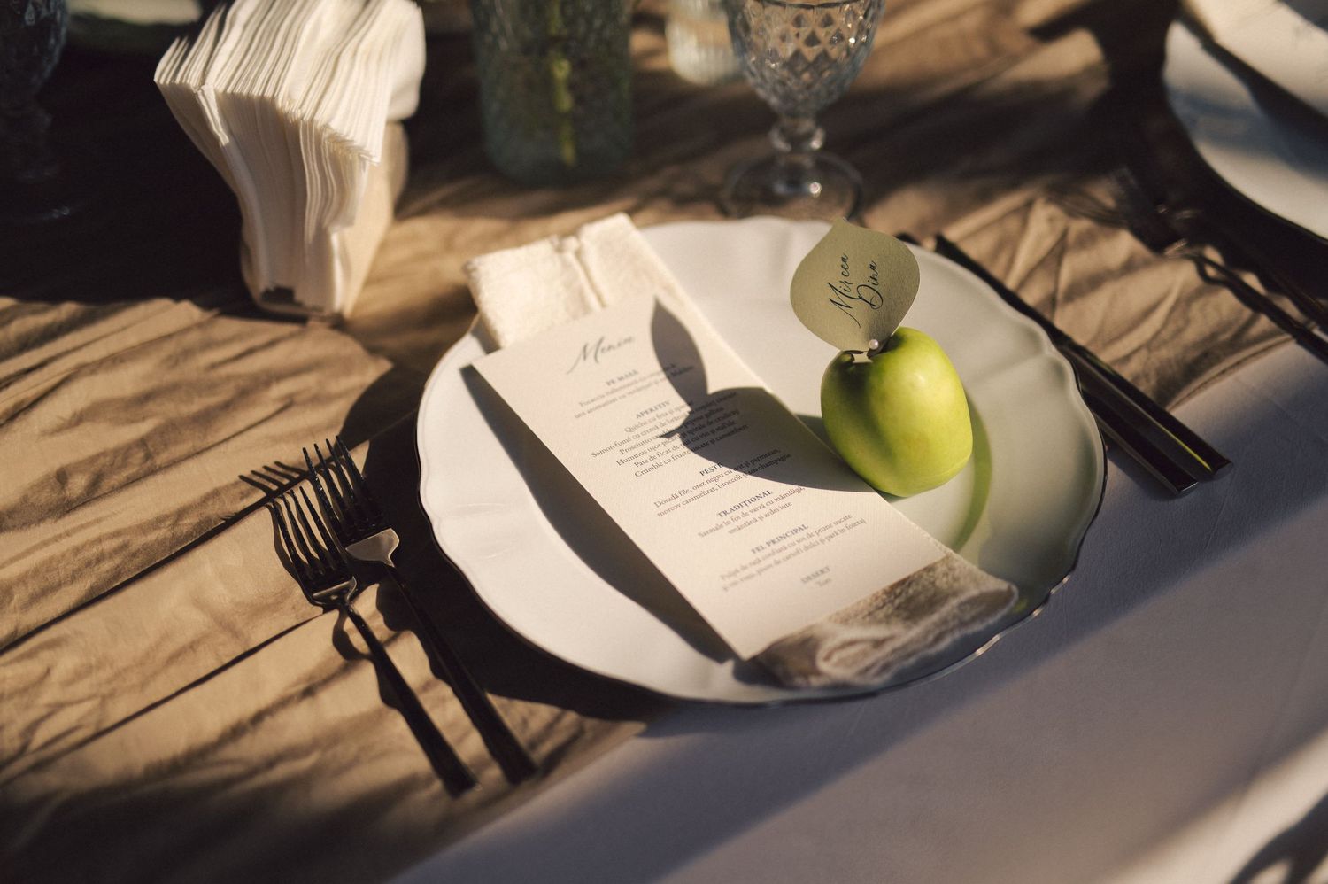 Place setting with white plate, utensils, green apple and crystal goblet on beige tablecloth in ambient lighting.
