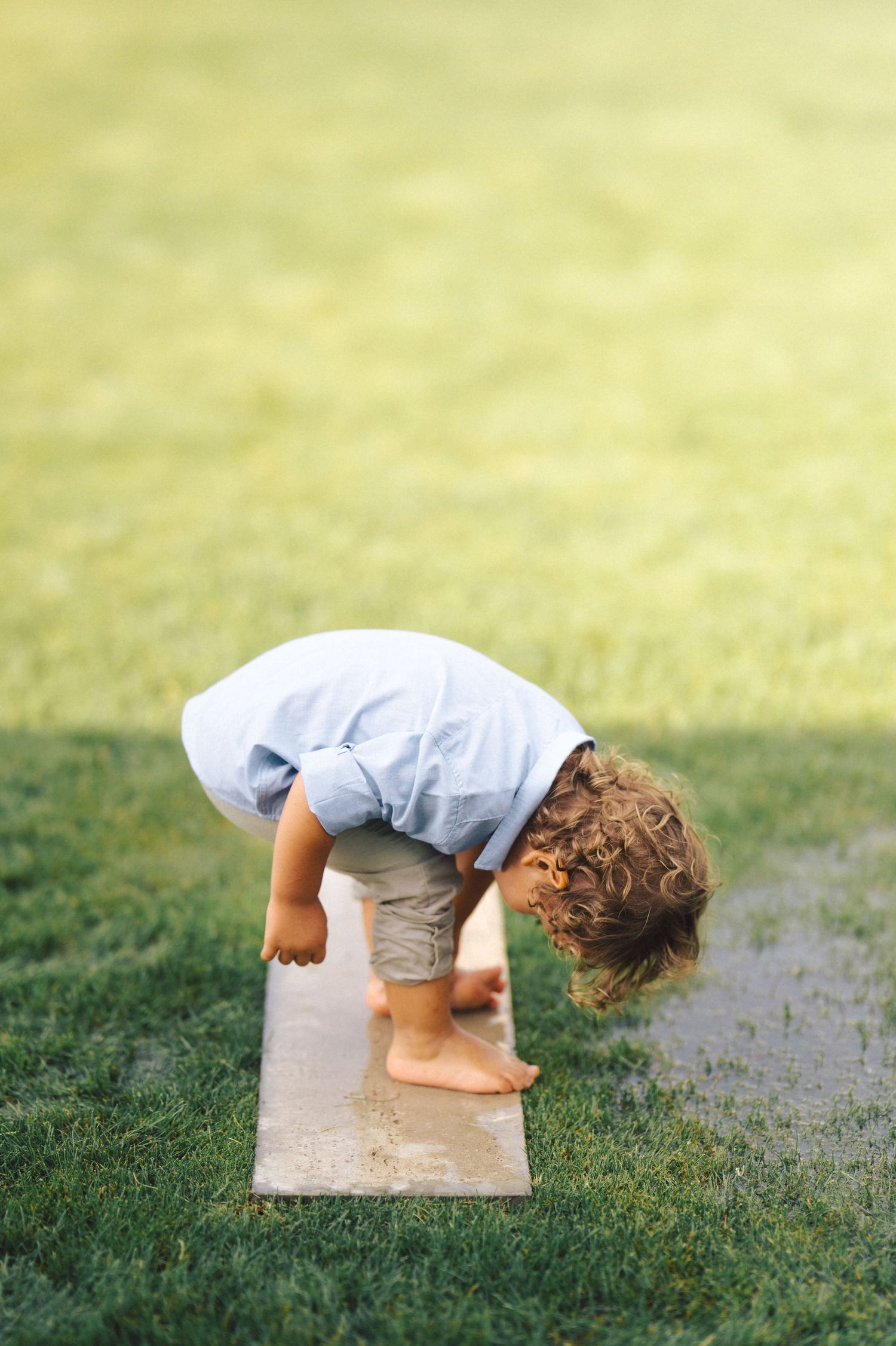 Small child bending over to look at something on a board in grass on a sunny day with bare feet.