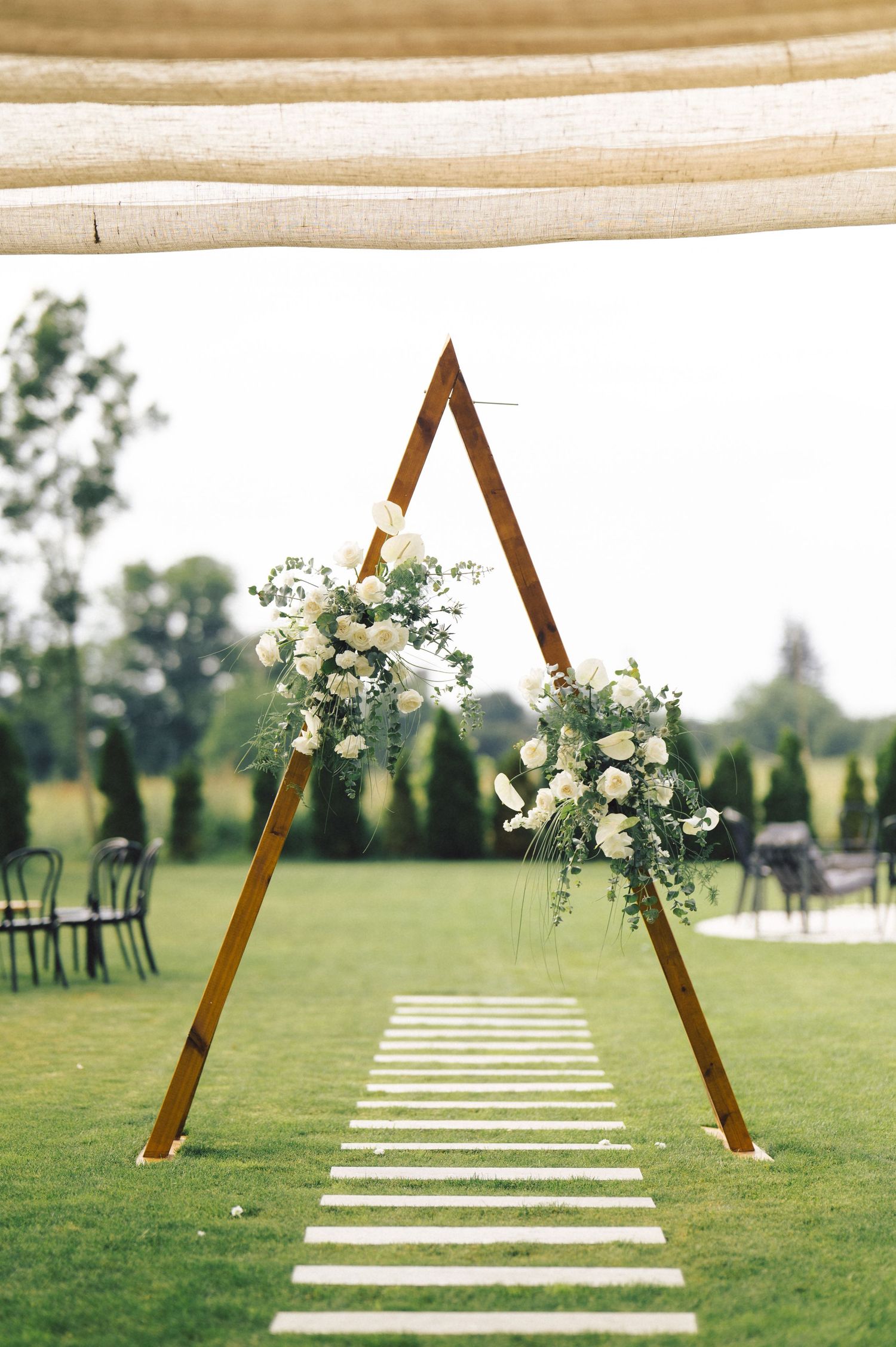 Wooden triangular wedding arch with white floral arrangements stands on green lawn with stone pathway leading to ceremony.