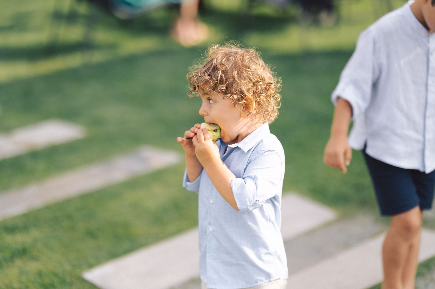 A child with curly hair in a white shirt enjoys eating outside in a sunny park while someone walks in the background.