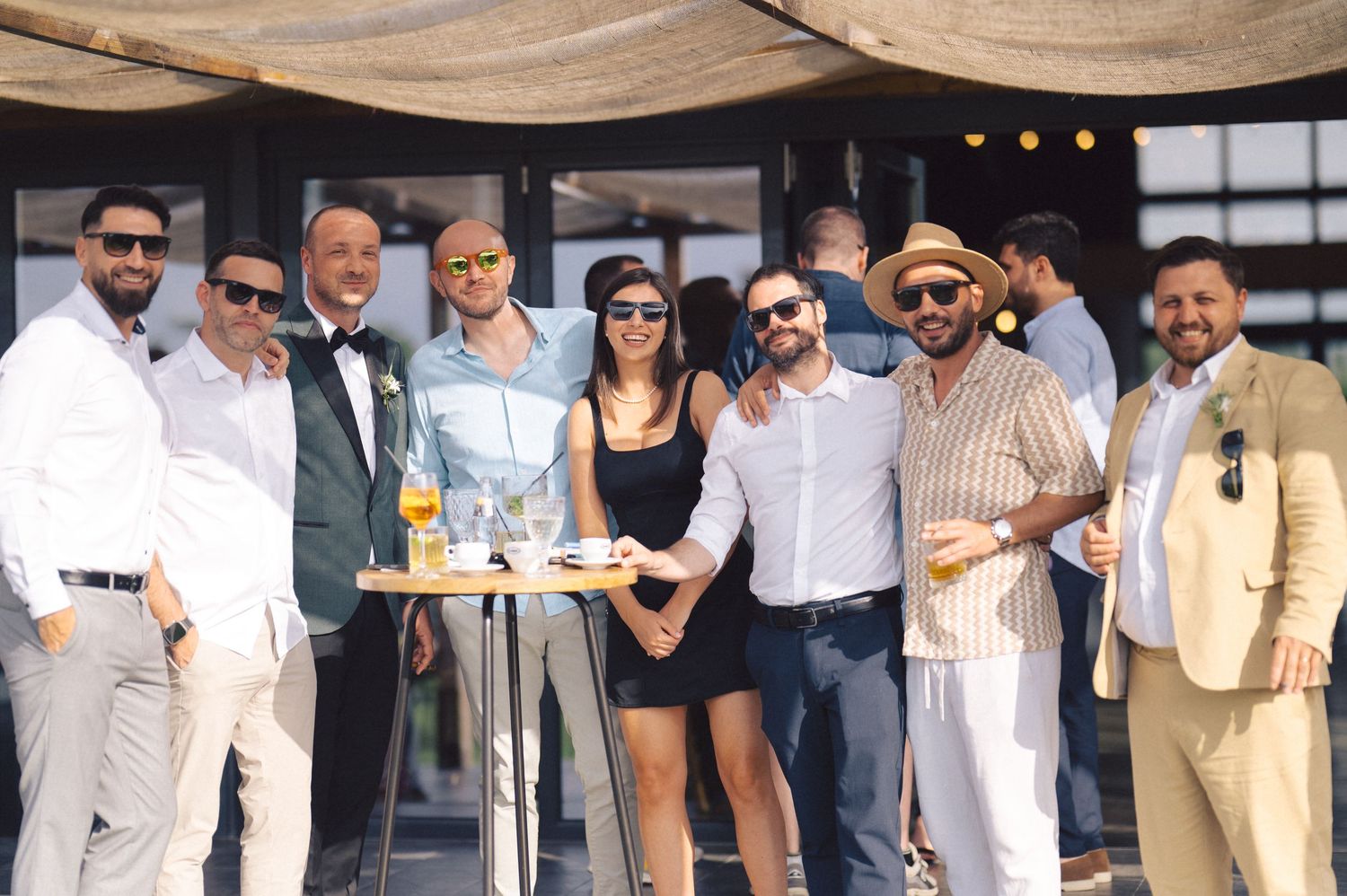 A stylishly dressed group poses together at an outdoor gathering with cocktails and sunglasses.