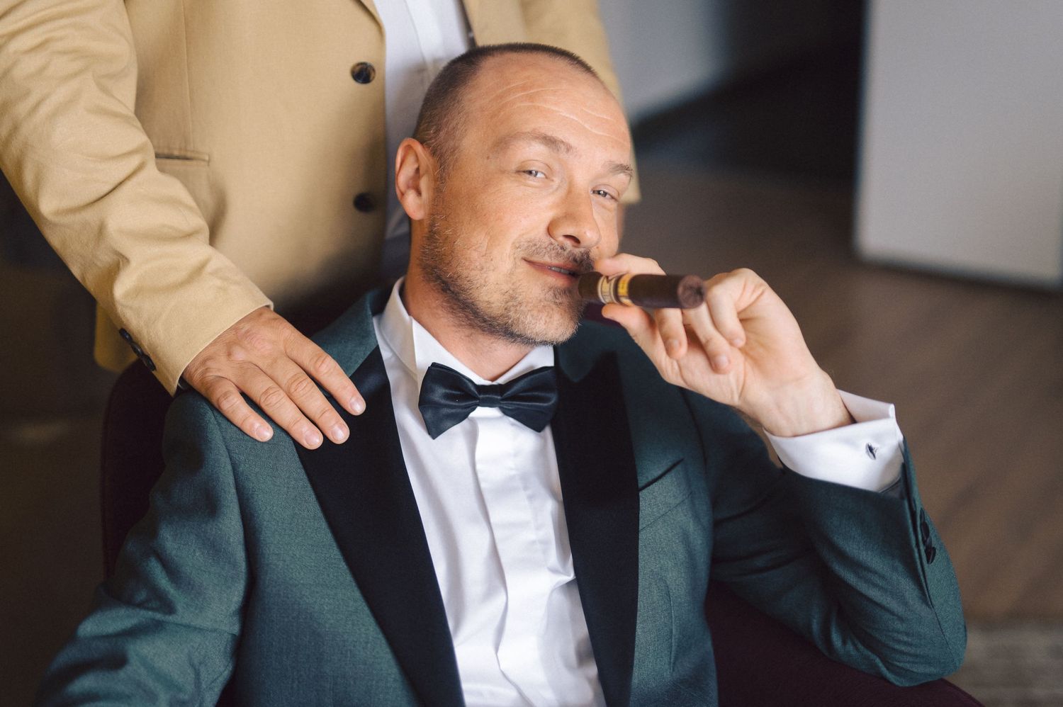 Person in emerald green suit jacket and bow tie smoking cigar while receiving shoulder massage.
