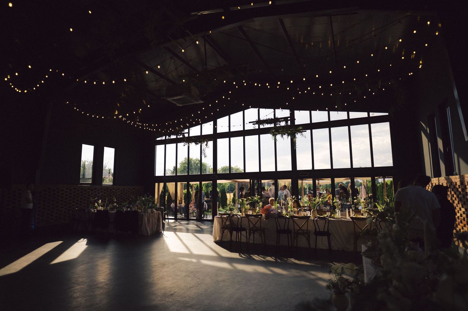 Interior of a dark reception venue with large windows, string lights, and sunlight streaming in creating dramatic shadows.