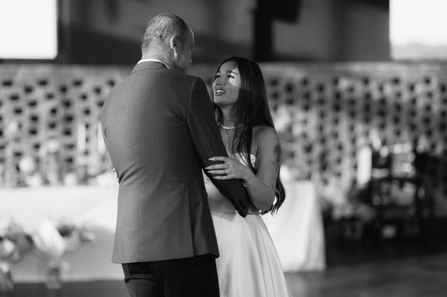 Black and white photo of a couple sharing their first dance at a wedding reception with stadium seating in background.