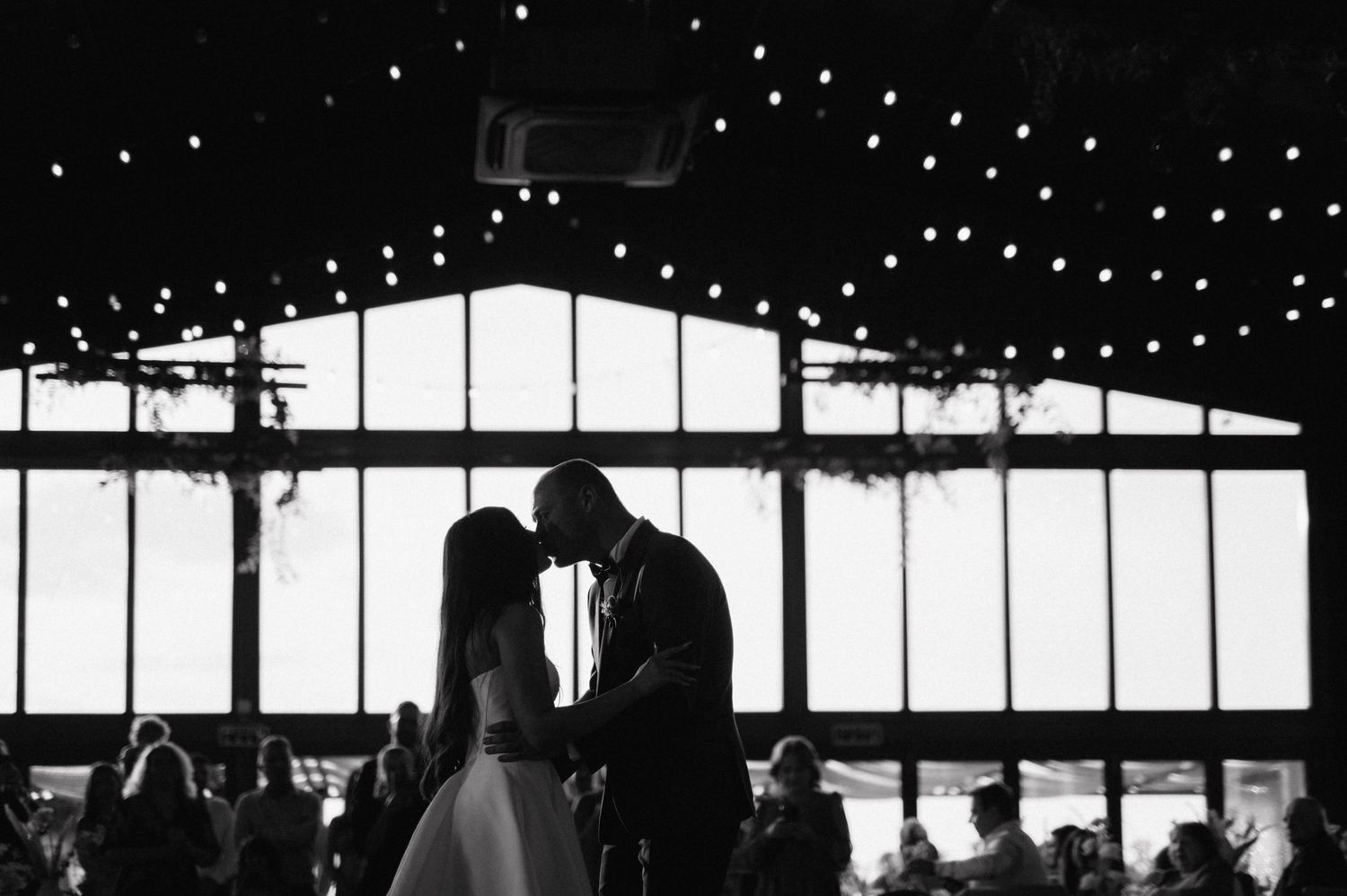 A romantic couple is silhouetted against glowing windows with string lights during an evening wedding reception.