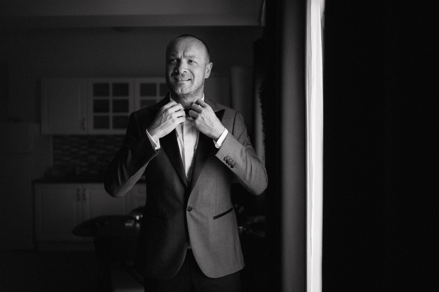 Black and white photo of someone adjusting their bow tie while wearing a formal suit and smiling.